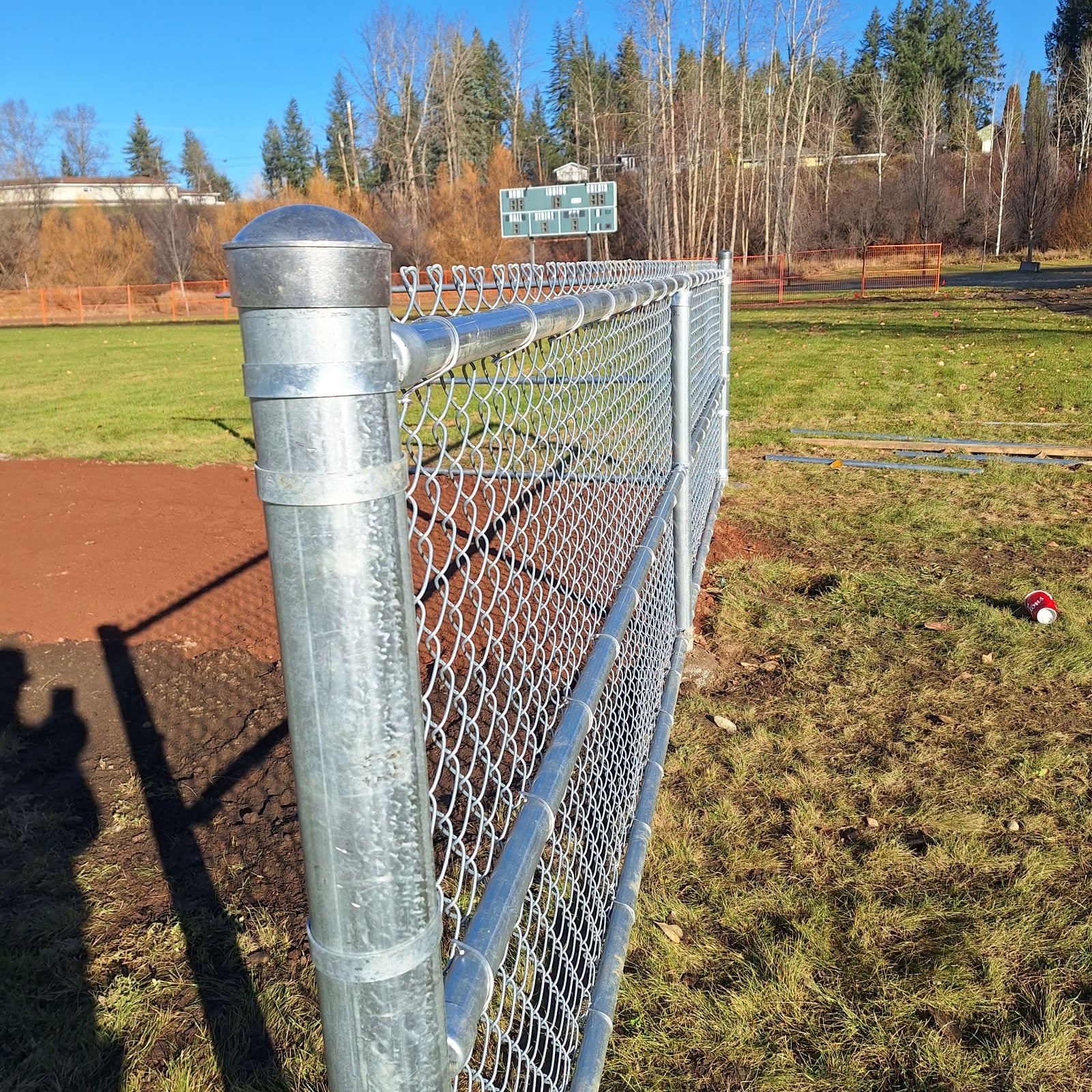 Metal chain-link fence around a sports field with a scoreboard in the background, trees, and clear blue sky.