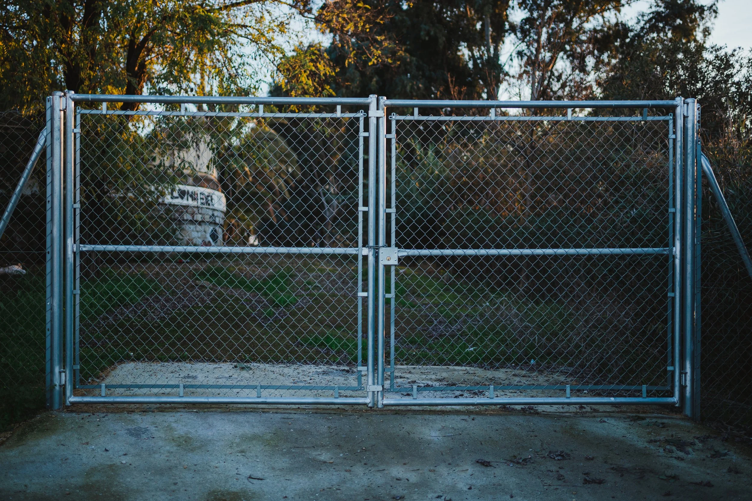 A metal chain-link gate with a padlock, set in a fenced area with trees and a sign that says 'LOVEE' in the background.