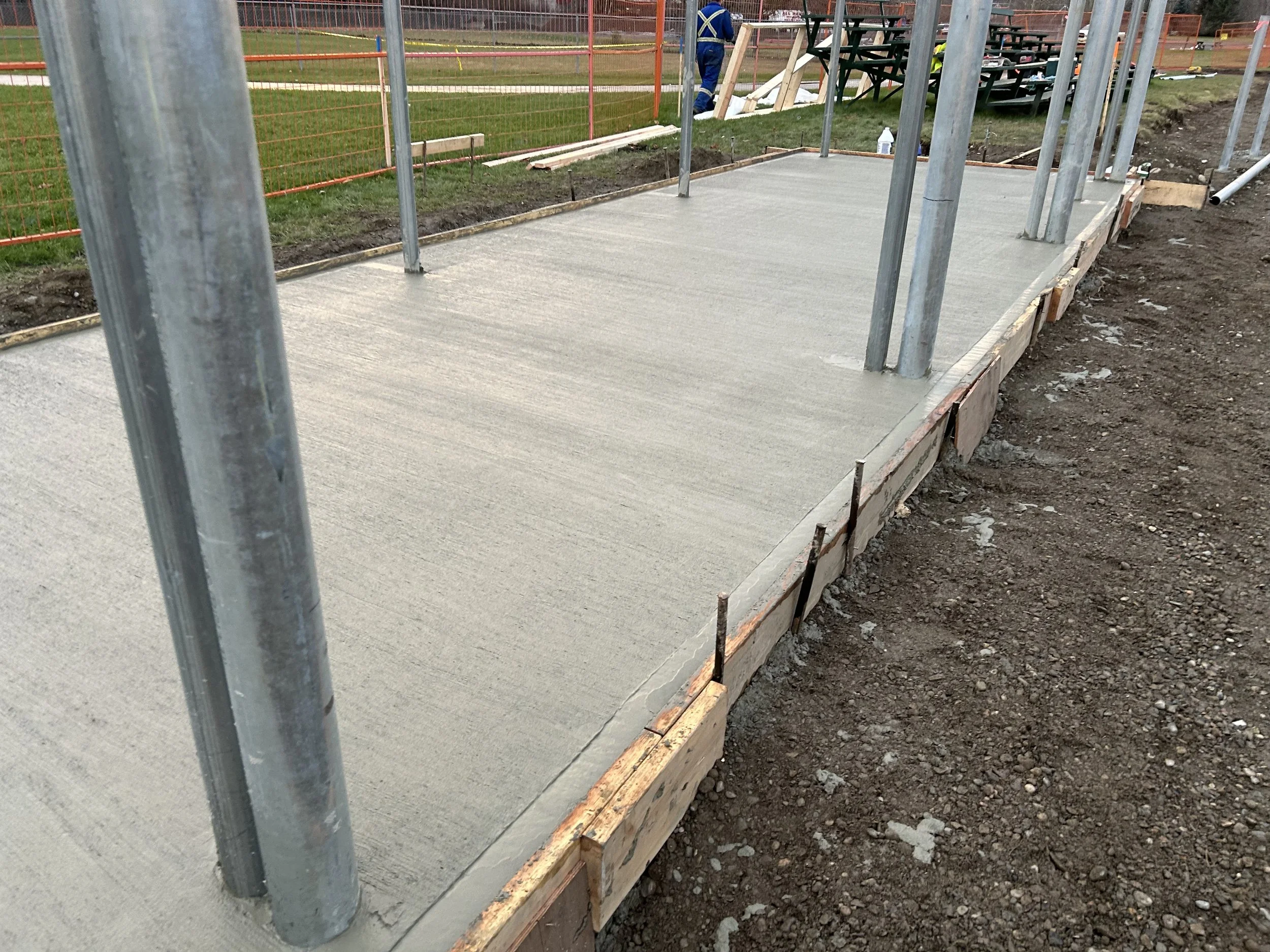 Freshly poured concrete on a construction site with metal poles for framing, wooden formwork, and gravel ground, and a worker in the background.