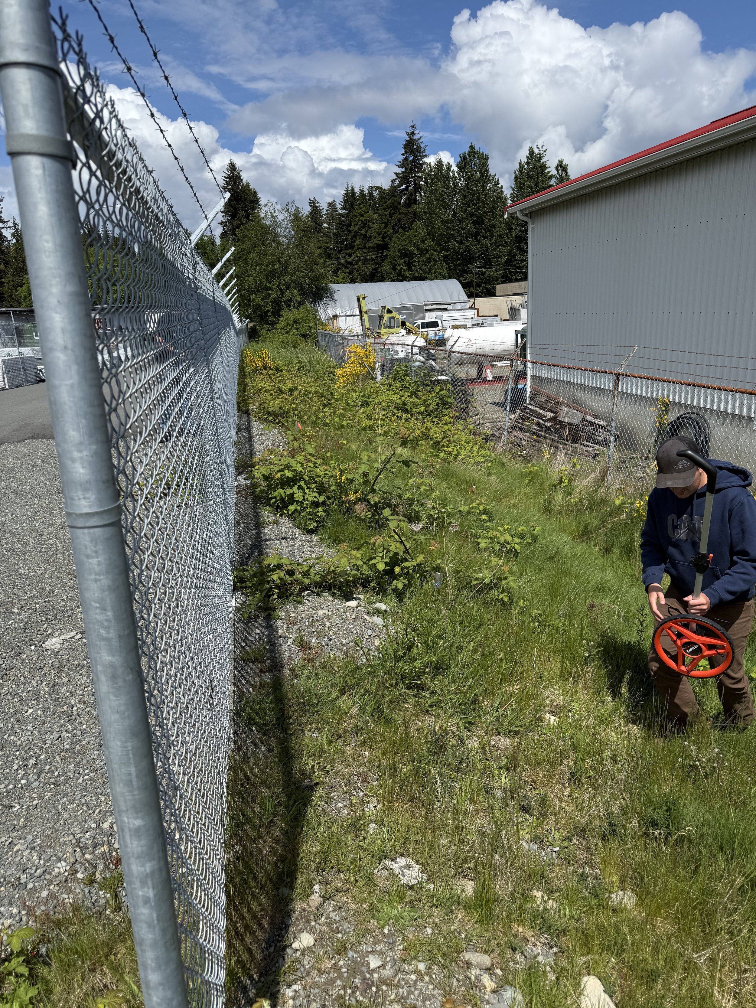 A person inspecting a chain-link fence with a screwdriver, surrounded by grass, bushes, and industrial buildings under a partly cloudy sky.