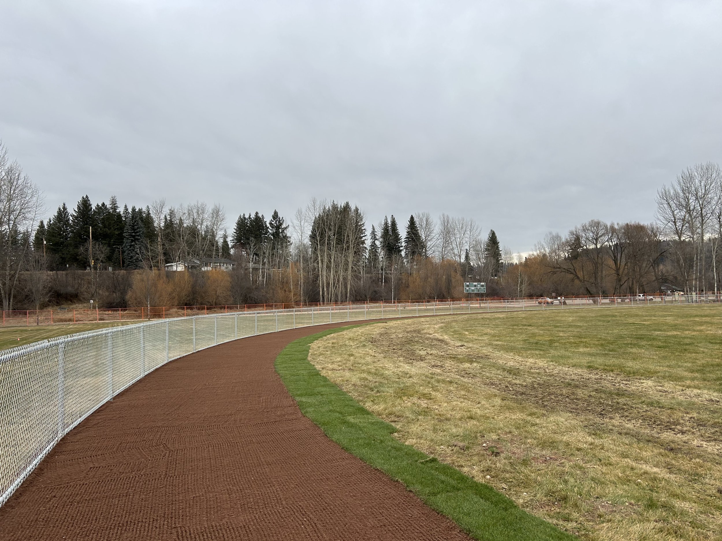 A view of a sports field with a dirt running track, a grassy area, a chain-link fence, and trees in the background under a cloudy sky.