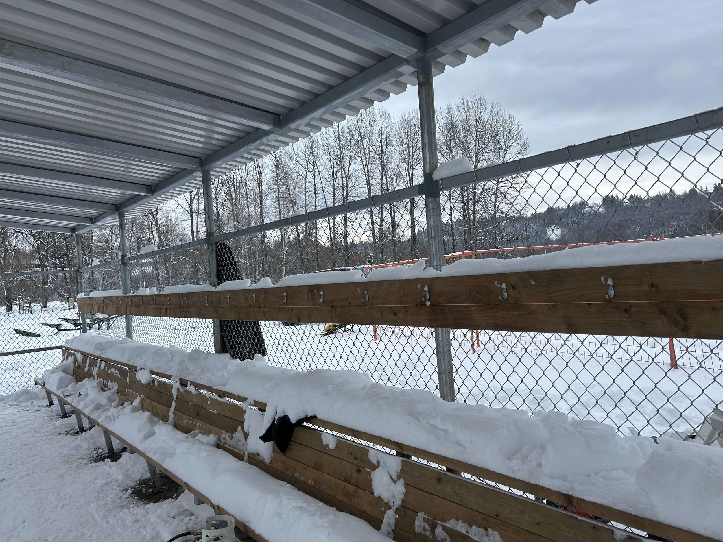 Empty outdoor sports bleacher seats covered with snow, located near a chain-link fence and surrounded by snow-covered ground and trees, under a cloudy sky.