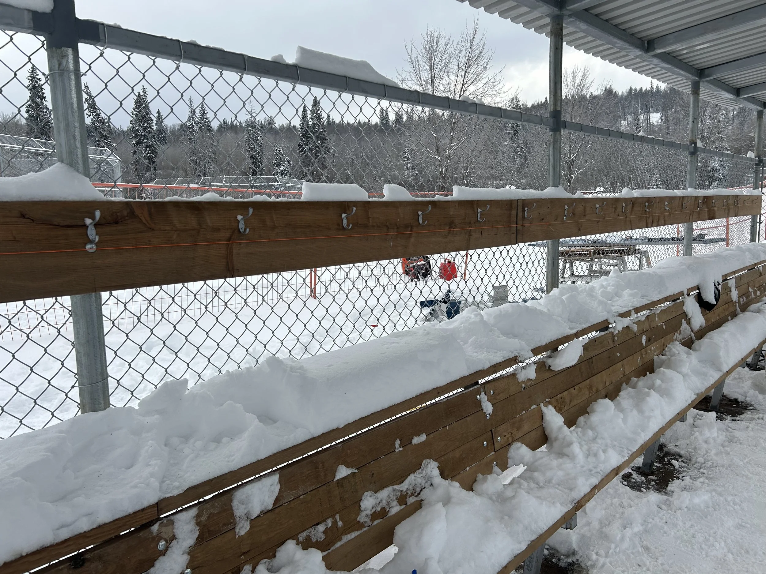 Snow-covered bleachers at an outdoor sports field with a chain-link fence and snowy landscape in the background.