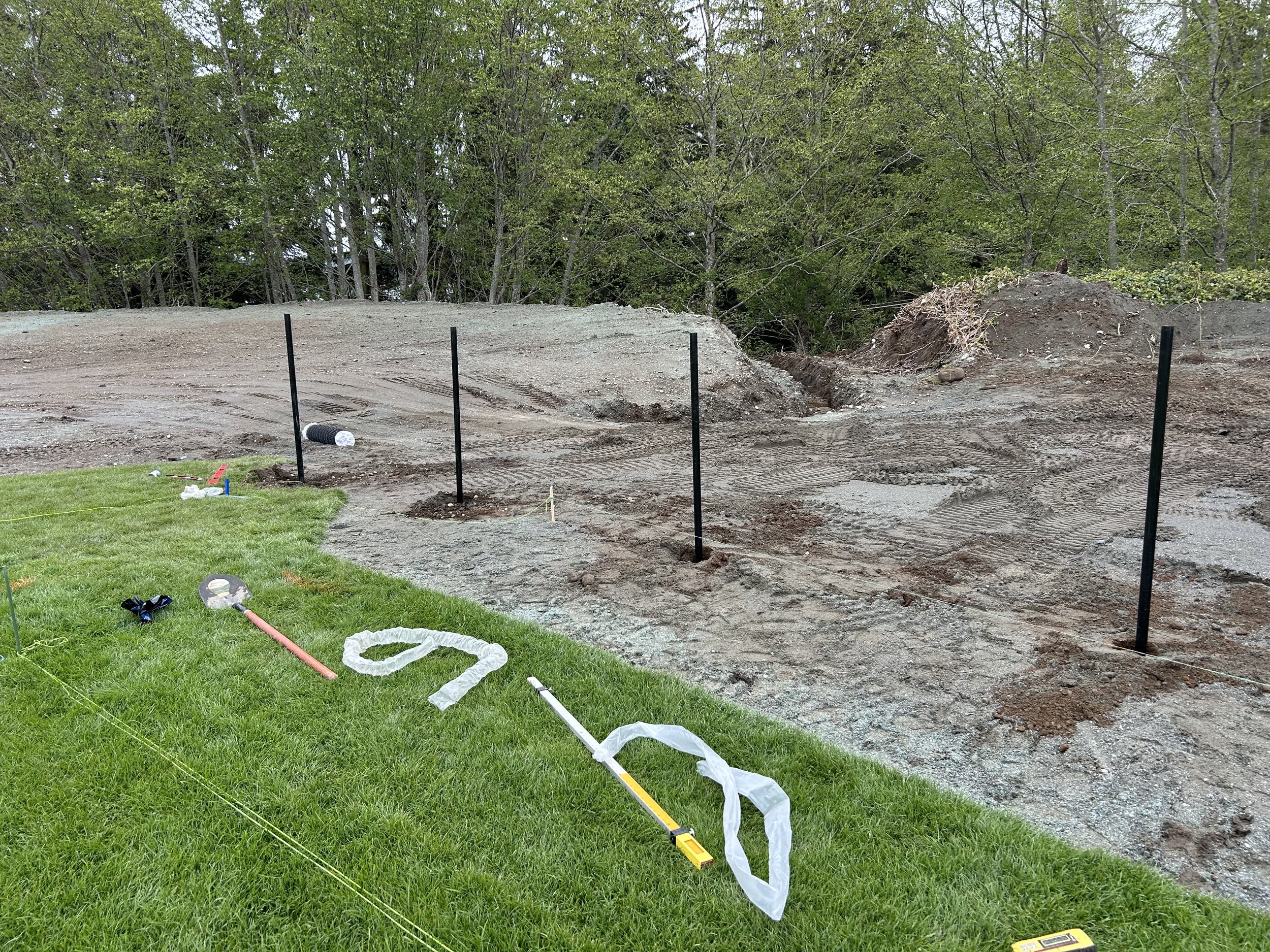 Construction site with black metal posts set in ground, disturbed earth, and a grassy area in the foreground with construction tools and markers.
