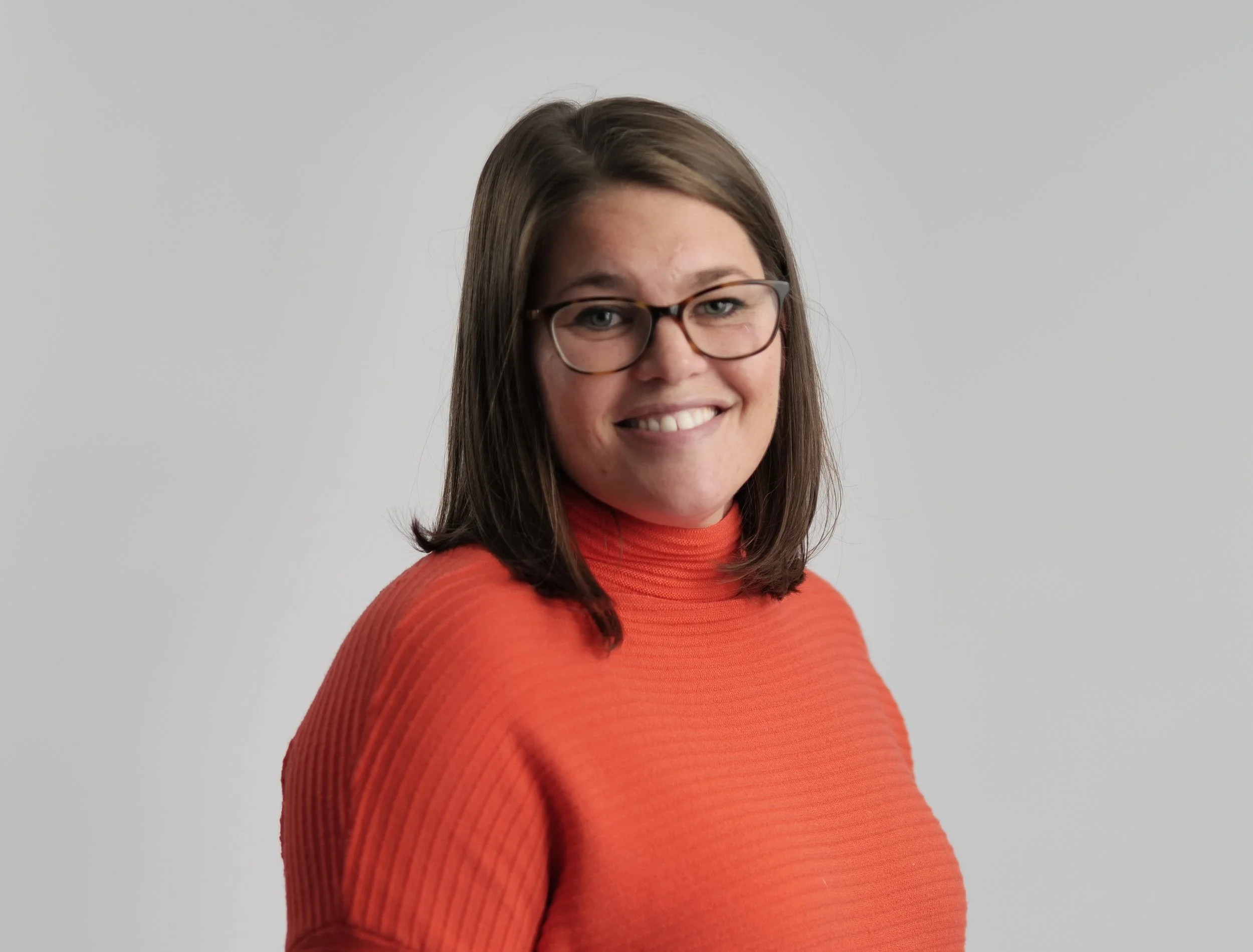A woman with shoulder-length brown hair, wearing glasses and a bright orange turtleneck sweater, smiling at the camera against a plain gray background.