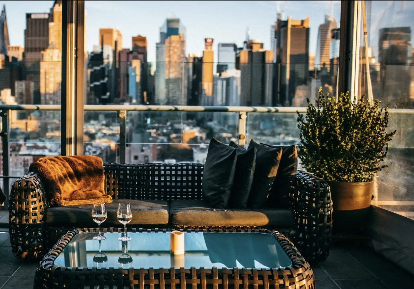 Outdoor balcony with wicker furniture, a glass-top coffee table with two glasses of wine, and a large potted plant, overlooking city skyscrapers during sunset.