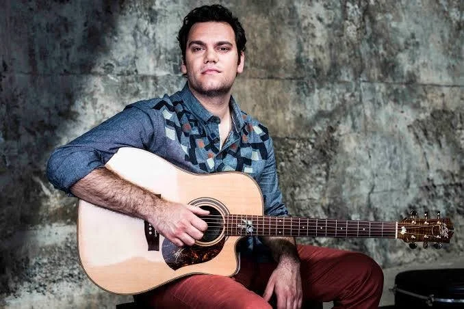 Man sitting with an acoustic guitar in front of a textured wall.