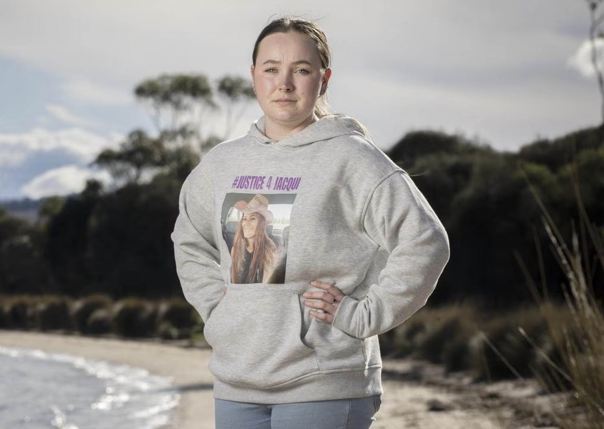 Young woman standing outdoors on a beach, wearing a gray hoodie with a photo and hashtag #JUSTICE4JACQUI printed on it. She has her hand on her hip, gazing into the camera with a serious expression.