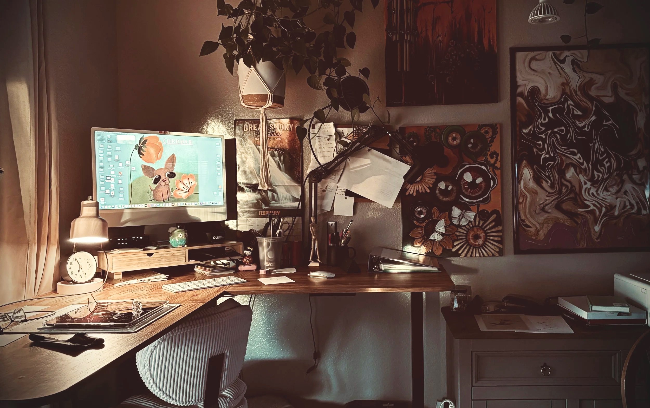 A cozy home office with a wooden desk, a computer monitor displaying a cartoon of a dog, a desk lamp, a clock, books, pens, and various artwork on the walls, including abstract paintings and flower designs.