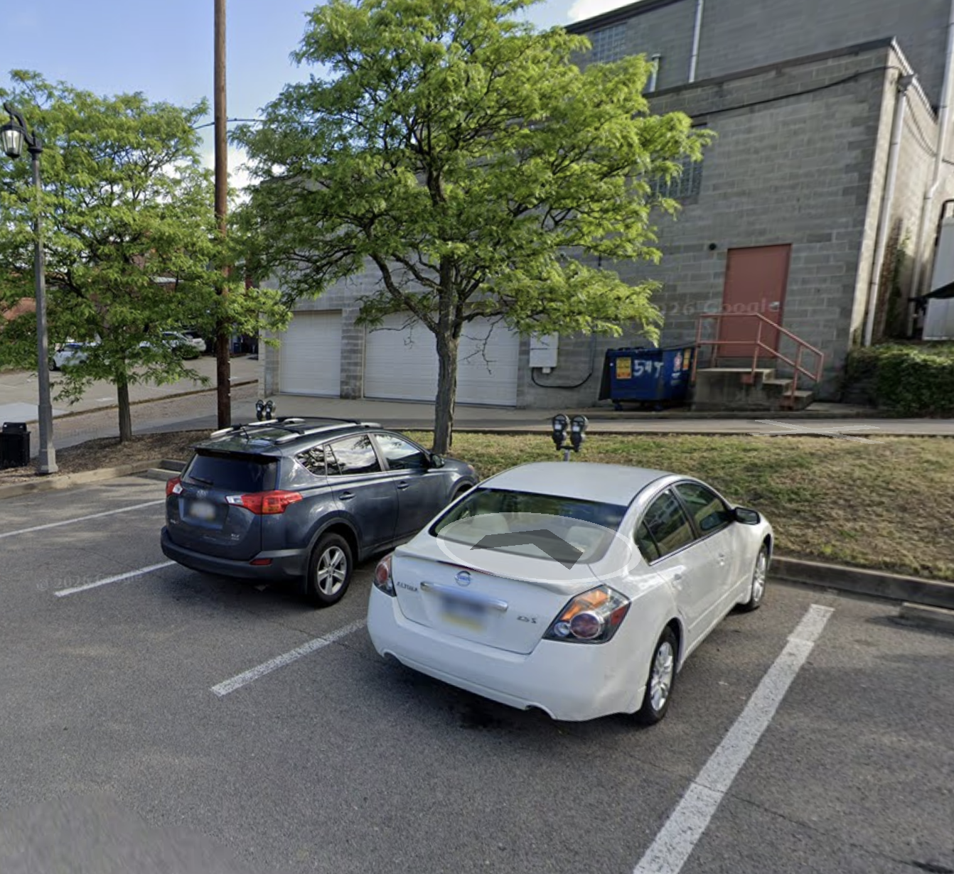 A parking lot with two cars, one black and one white, parked next to each other. There are two trees and a grassy area behind the cars. In the background, there are garage doors, a large building, a dumpster, and a sidewalk with steps leading up to a red door.