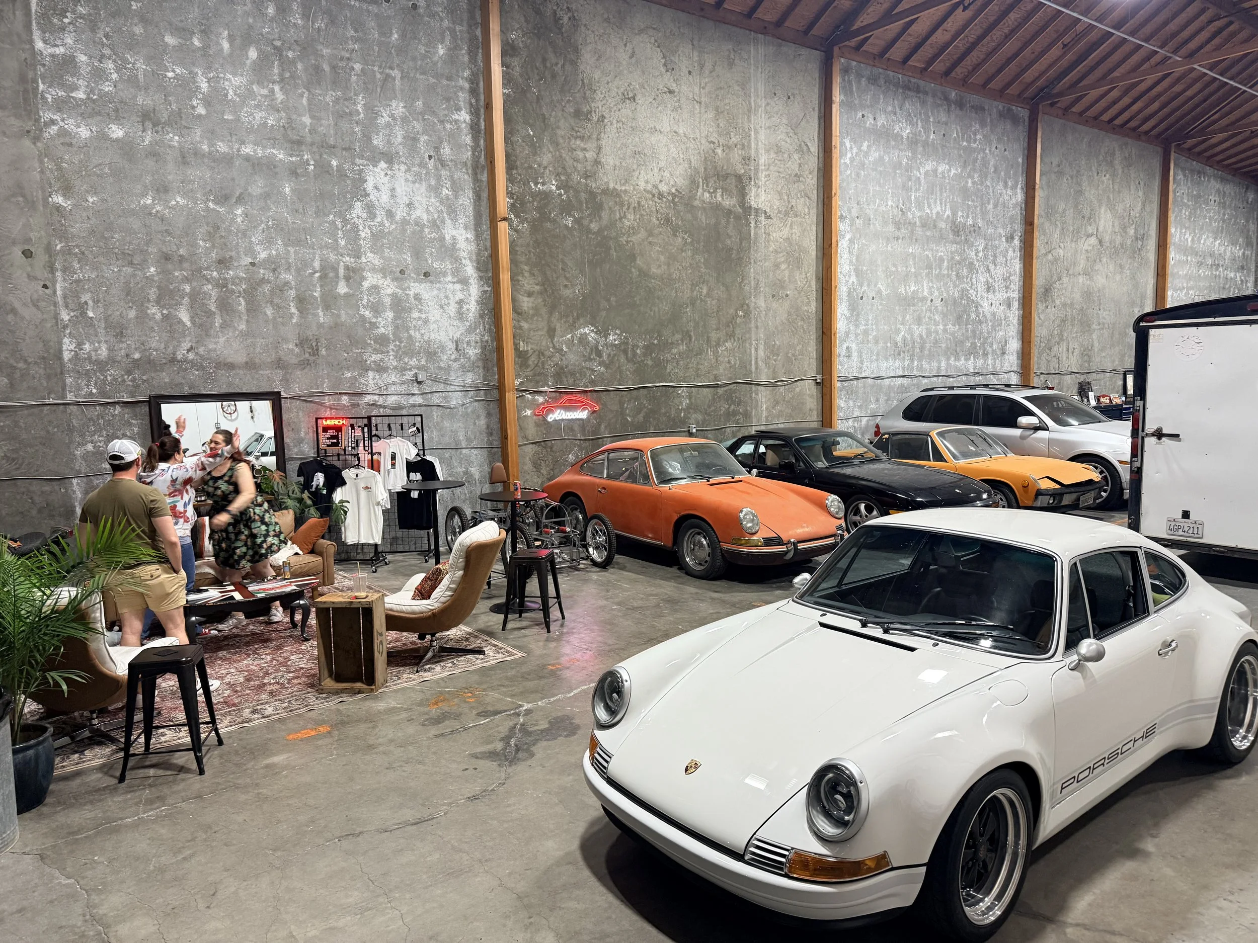 Interior of a vintage car showroom or gathering space with several classic and modern Porsche cars, including a white Porsche 911 in the foreground, an orange Porsche 911, and a black Porsche 911 in the background. There is a seating area with chairs, a small table, and a group of people chatting near a clothing display and mirror on the left side of the image.