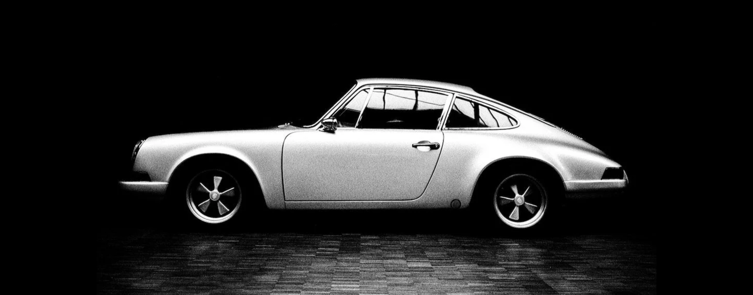A black and white photograph of a vintage sports car, possibly a Porsche, displayed on a reflective floor with a dark background.