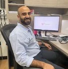 A man with a beard and short hair, wearing a light blue shirt, sitting at a desk in front of a computer monitor in an office setting.