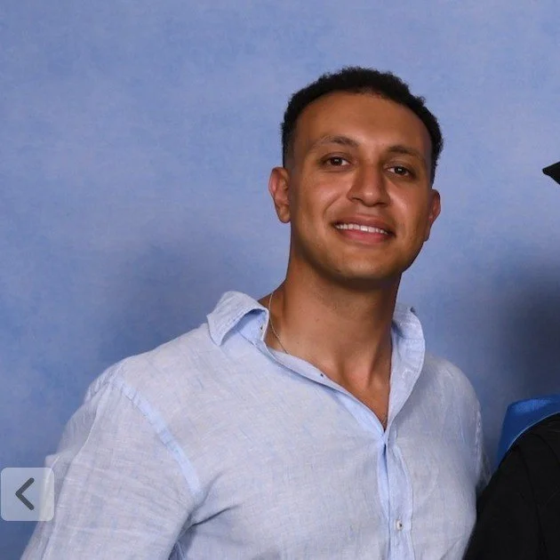 A young man smiling, wearing a light blue button-down shirt, standing against a plain blue background.