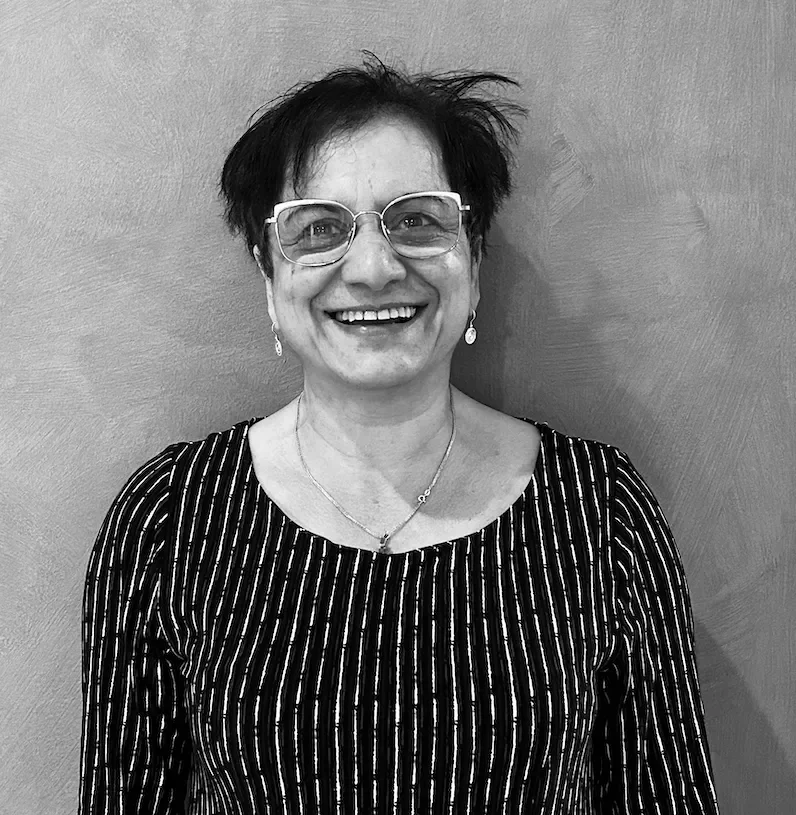 A woman with short black hair, glasses, and earrings smiling, wearing a striped top and a necklace, standing against a plain wall.