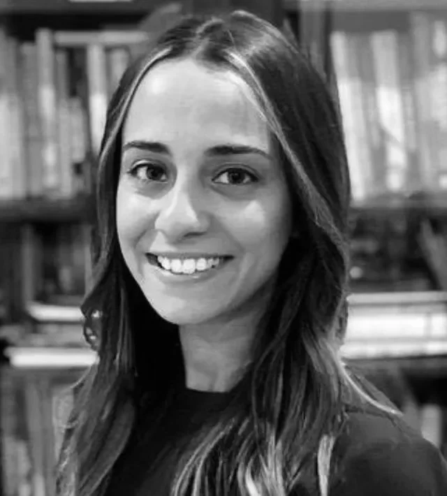 A young woman with long dark hair smiling in front of a bookshelf.