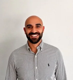 A man with a beard and short hair, wearing a light blue shirt, sitting at a desk in front of a computer monitor in an office setting.
