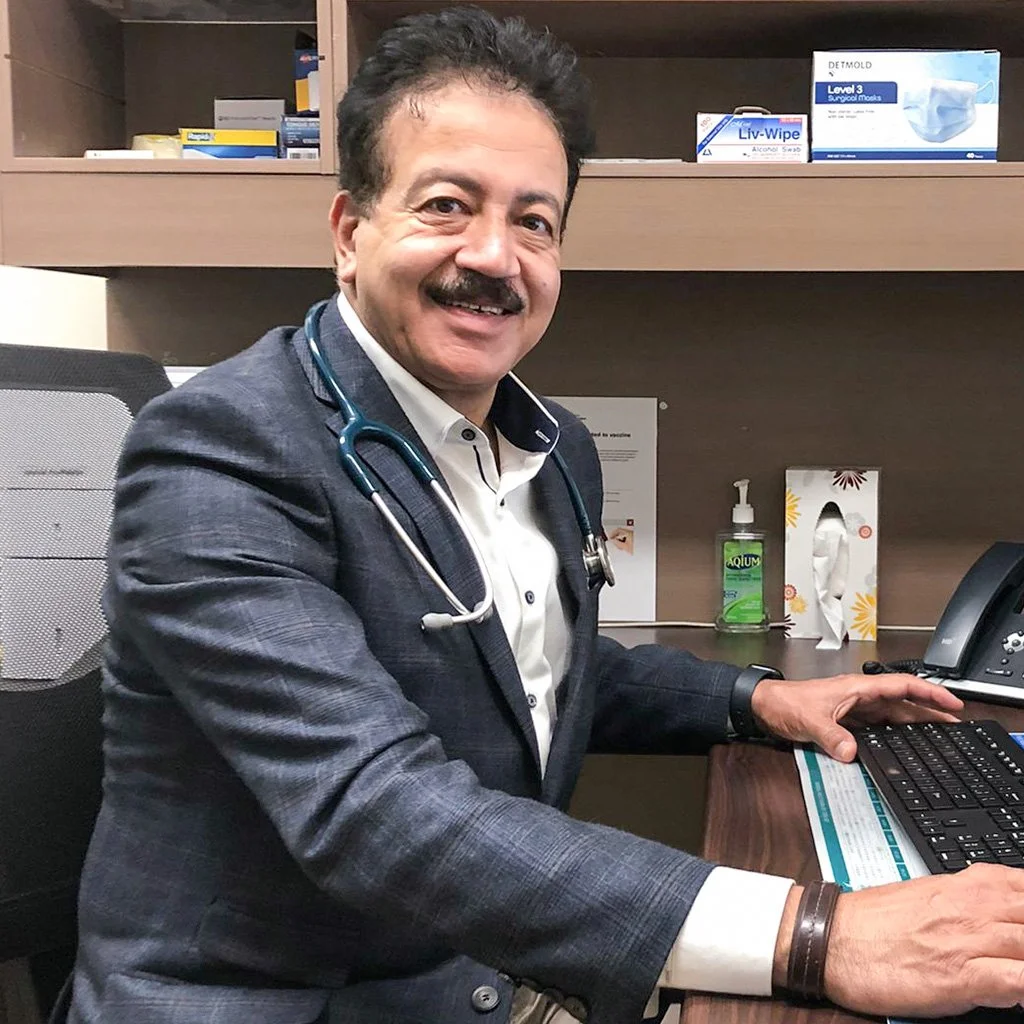 A male doctor wearing a gray suit with a white shirt and stethoscope around his neck, sitting at a desk in an office, smiling at the camera.