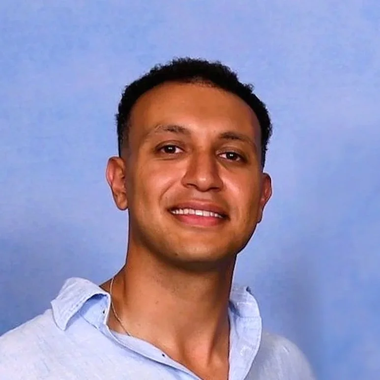 A young man smiling, wearing a light blue button-down shirt, standing against a plain blue background.