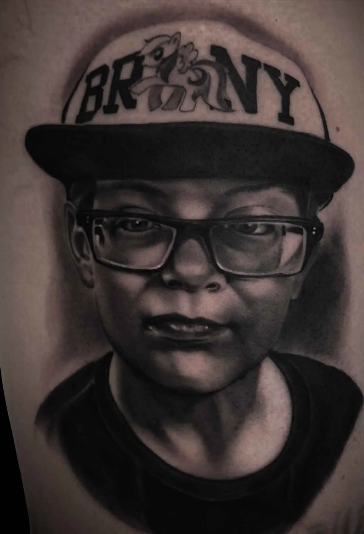 a black and grey portrait tattoo of a young boy wearing a brony hat