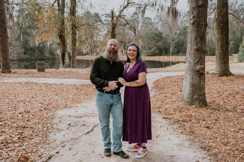 A smiling man with a beard and a woman with colorful hair standing on a leaf-covered park path surrounded by large trees in autumn, near a small lake.