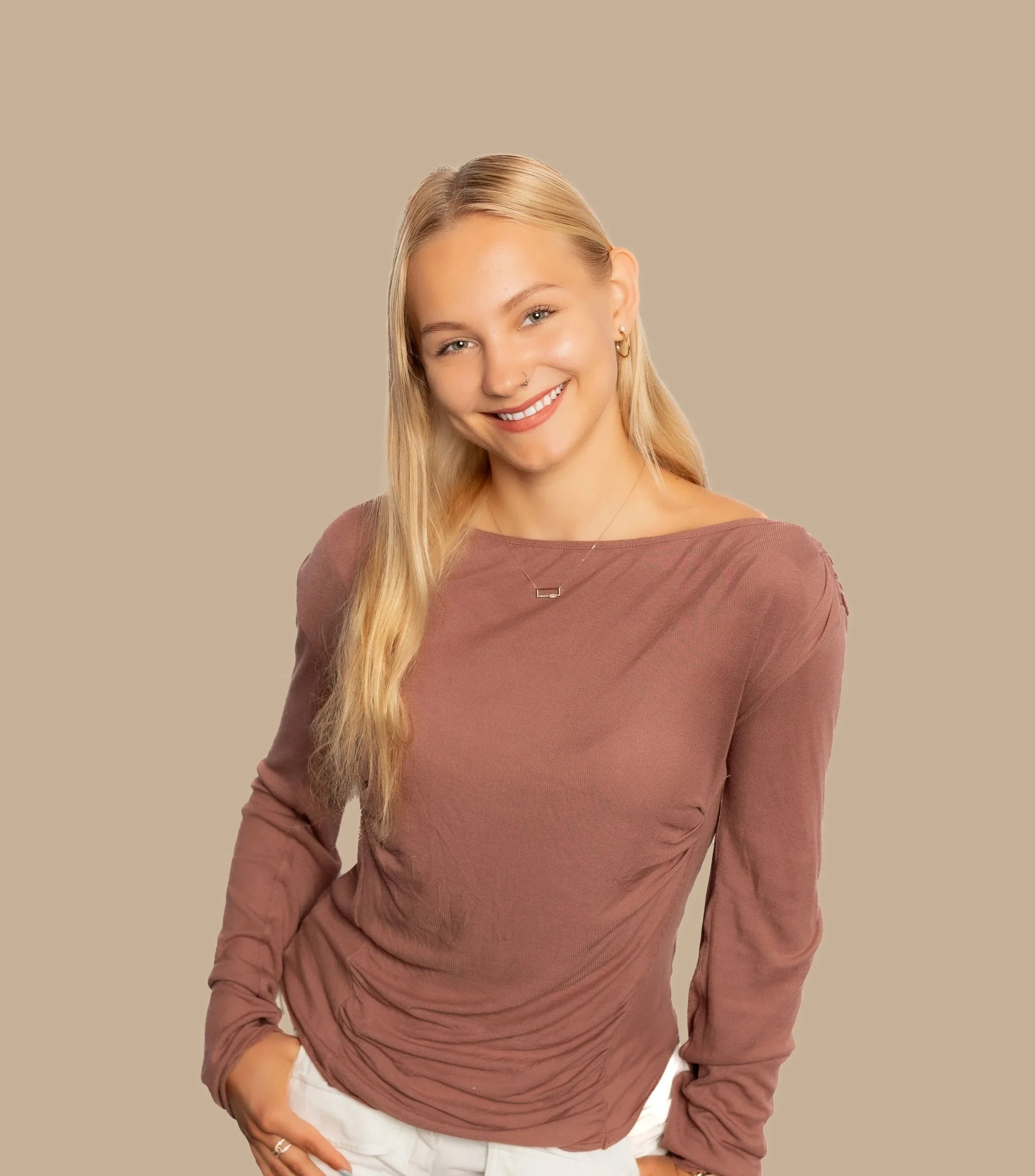 Portrait of a young woman with blonde hair, smiling, wearing a mauve long-sleeve top and white pants, against a beige background.
