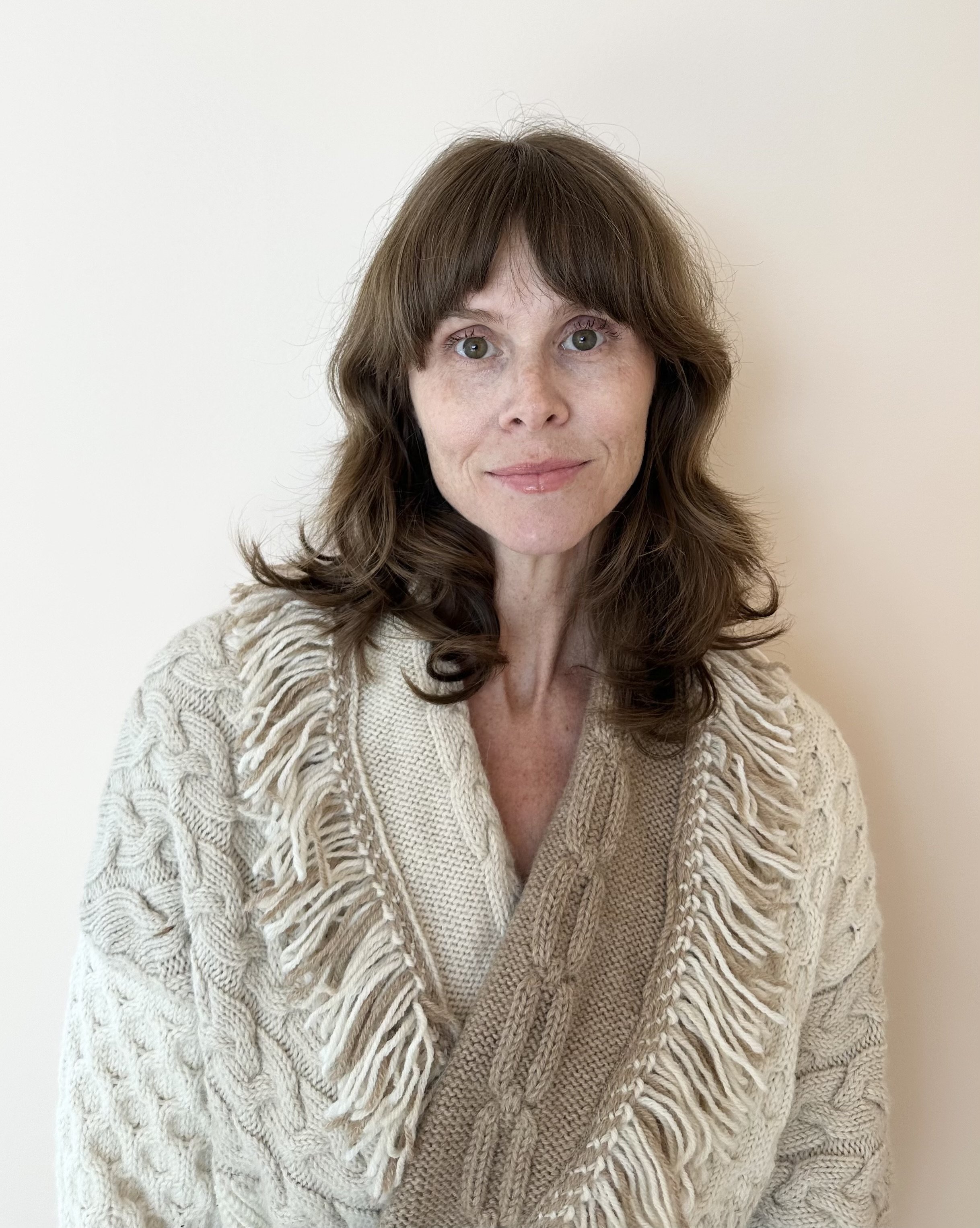 A woman with wavy brown hair wearing a beige cable-knit sweater standing against a plain white wall.
