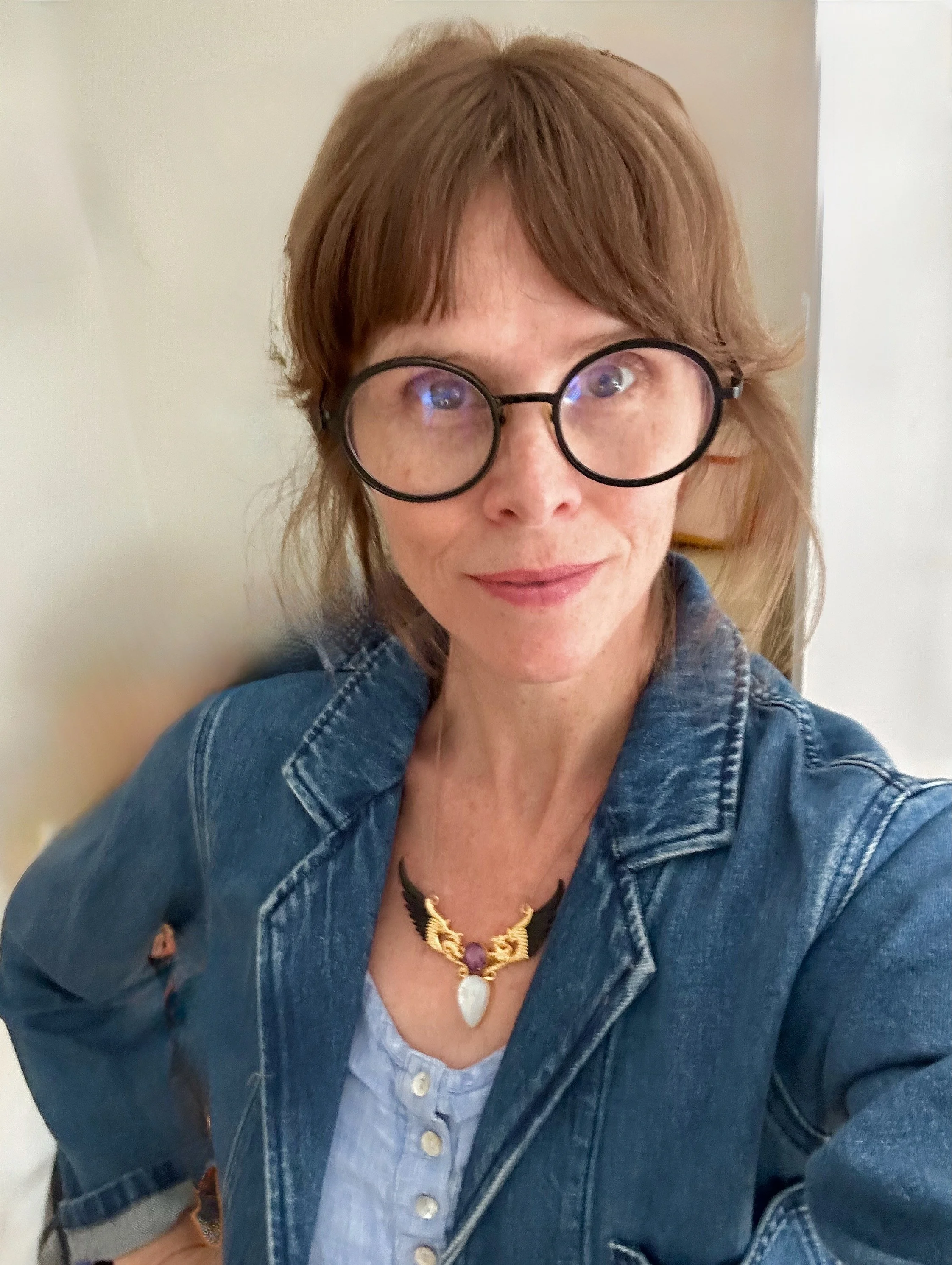 A woman with red hair wearing large round glasses, a denim jacket, a light blue button-up shirt, and a jewelry necklace, in a room with a plain background.