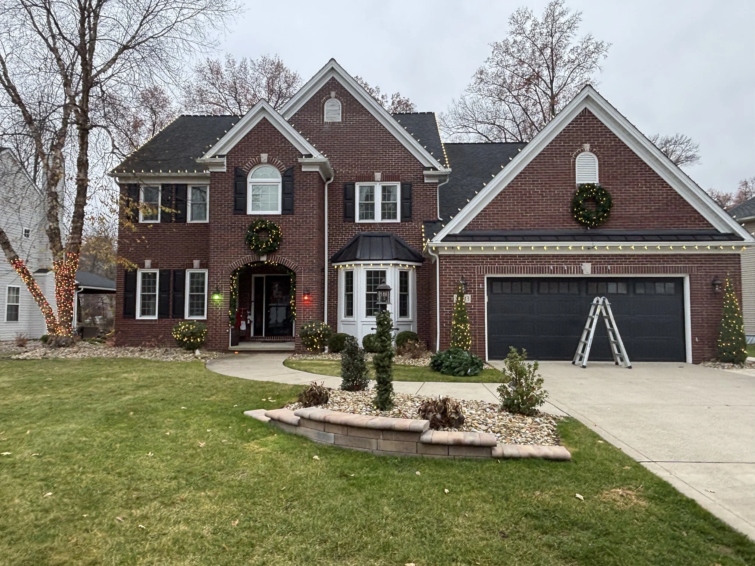 Red brick two-story house decorated with Christmas wreaths and string lights, with a lawn and garden in front, a ladder near the garage, and trees with Christmas lights in the background.