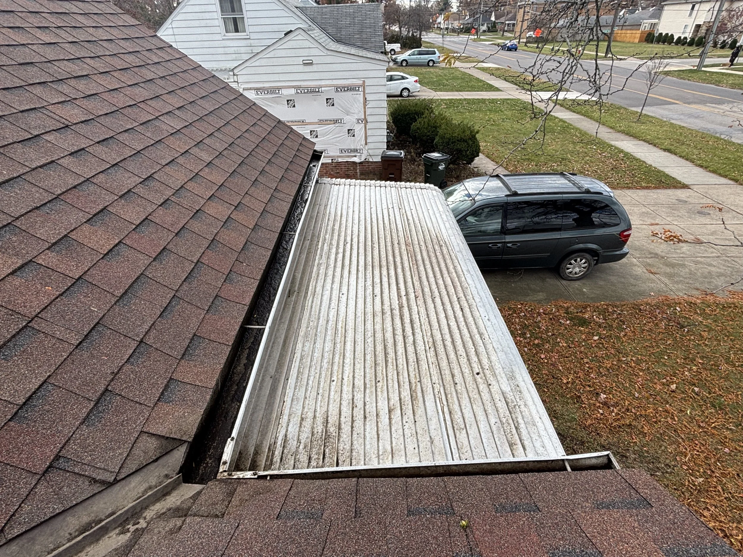 A residential rooftop with asphalt shingles, a flat metal roof section, and view of a neighborhood street with parked cars, sidewalk, trees, and houses in the background.