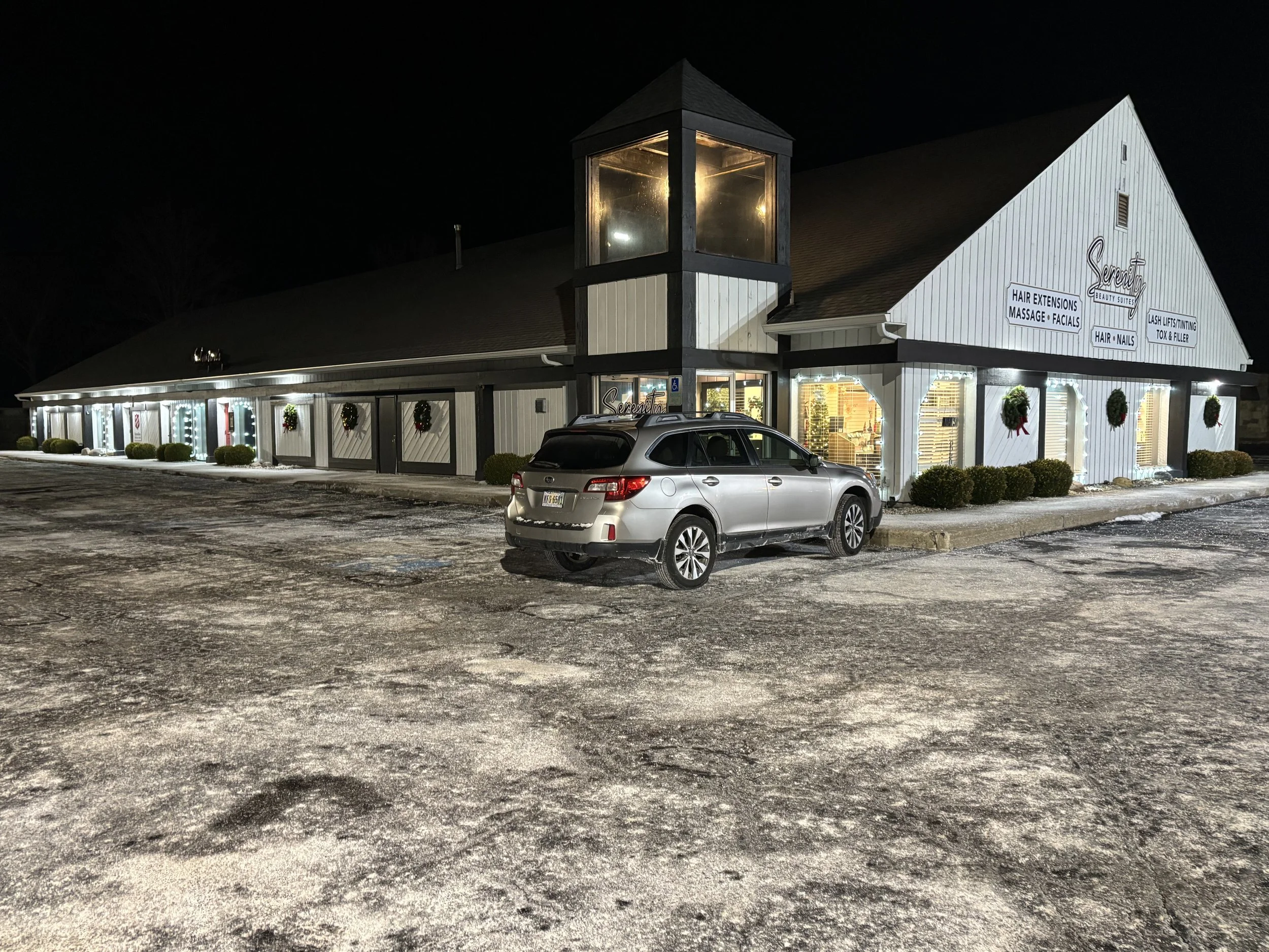 Night view of a white building with signs for a beauty salon, decorated with wreaths and wreath-style decorations, and a silver car parked in front in a snow-dusted parking lot.