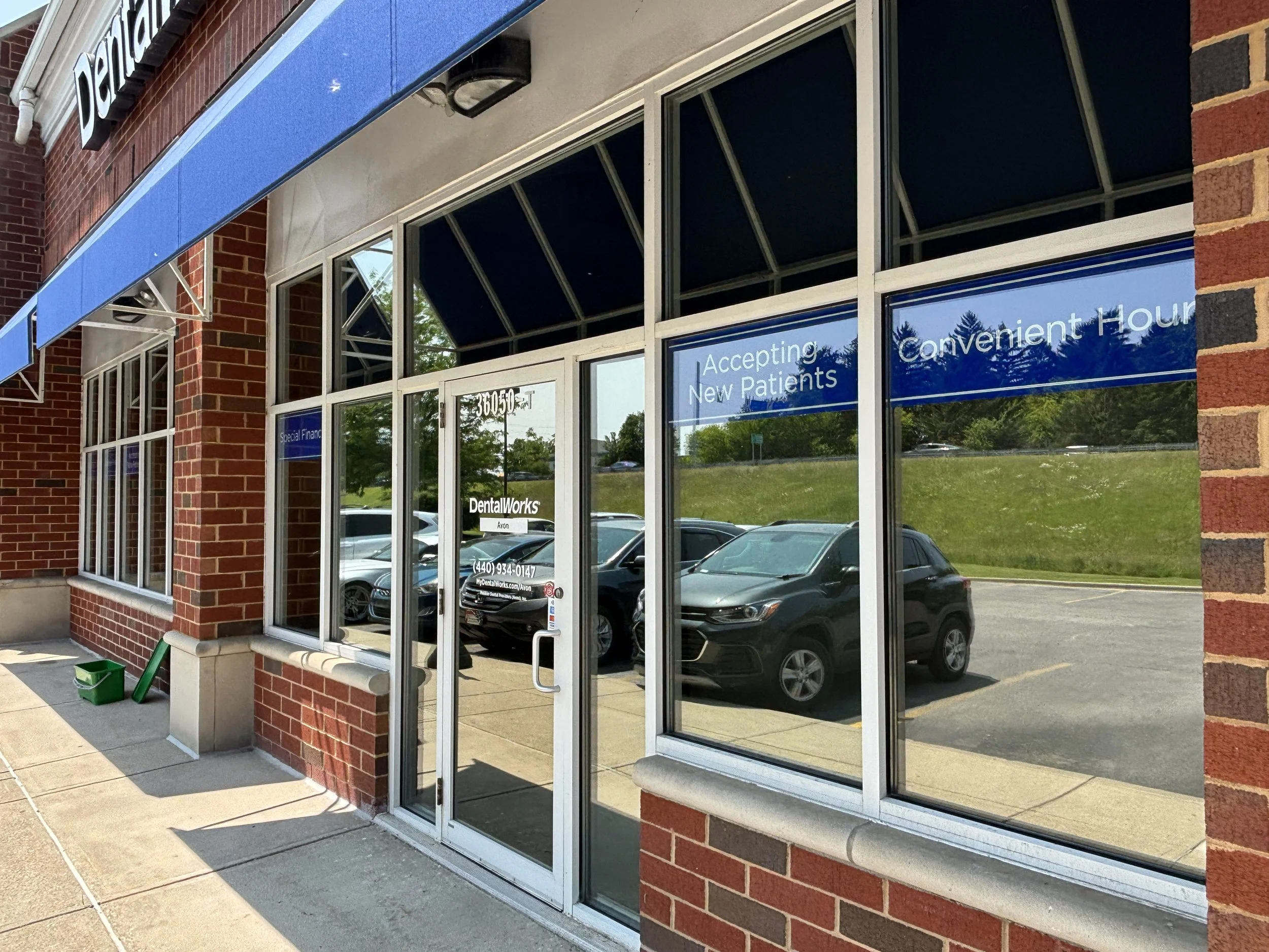 Exterior view of a dental office with large windows and blue awning, displaying signs that indicate accepting new patients and convenient hours, with parked cars reflected in the glass.