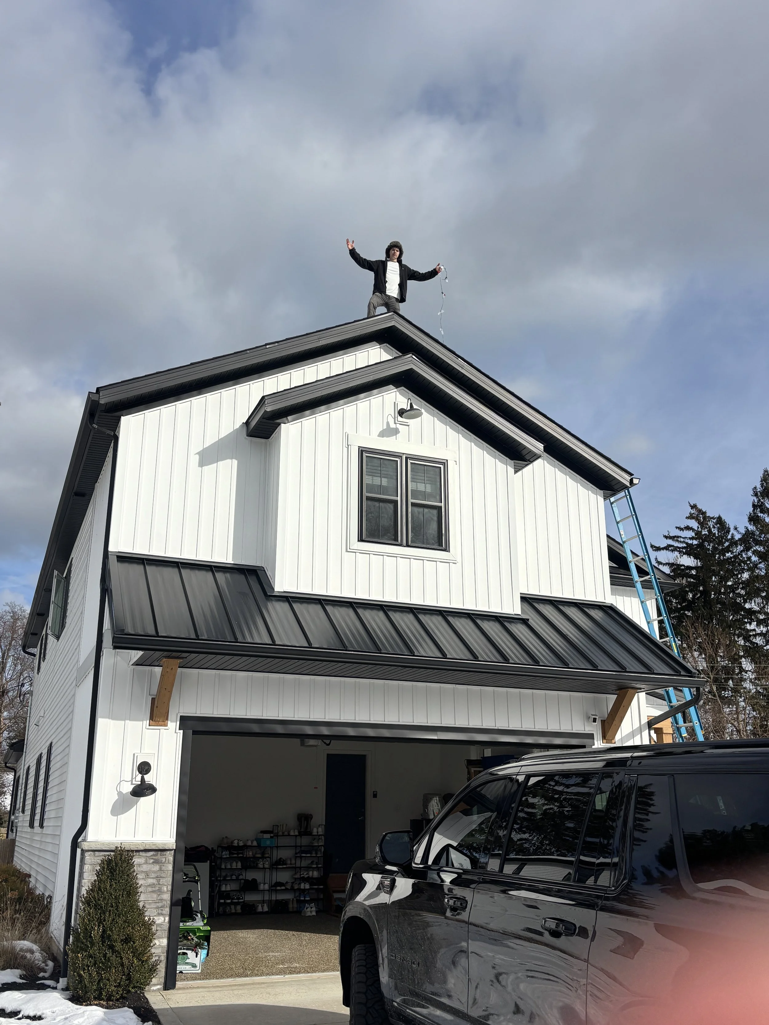 Person standing on the roof of a two-story house, waving with one arm and holding a string of lights in the other, with a ladder leaning against the house and a black vehicle parked in the driveway.