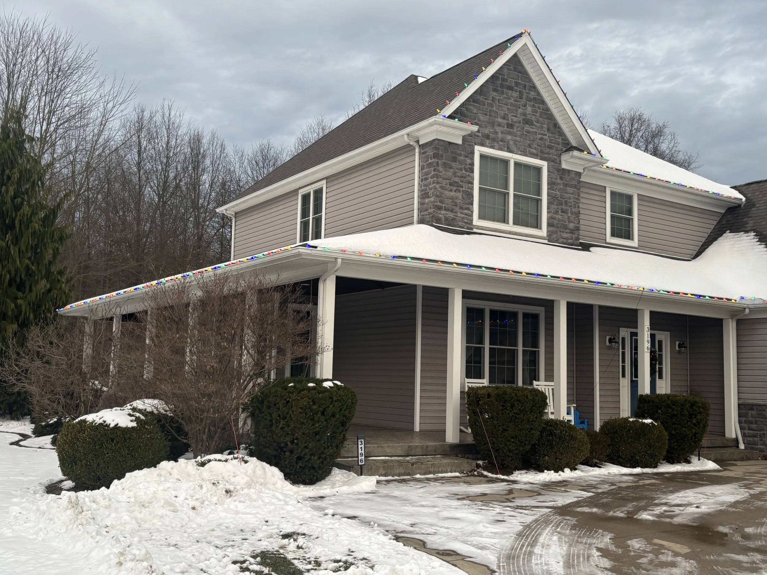 Two-story house with a snow-covered roof, decorated with multicolored Christmas lights along the roofline, and surrounded by snow and bushes, with a driveway in the foreground.