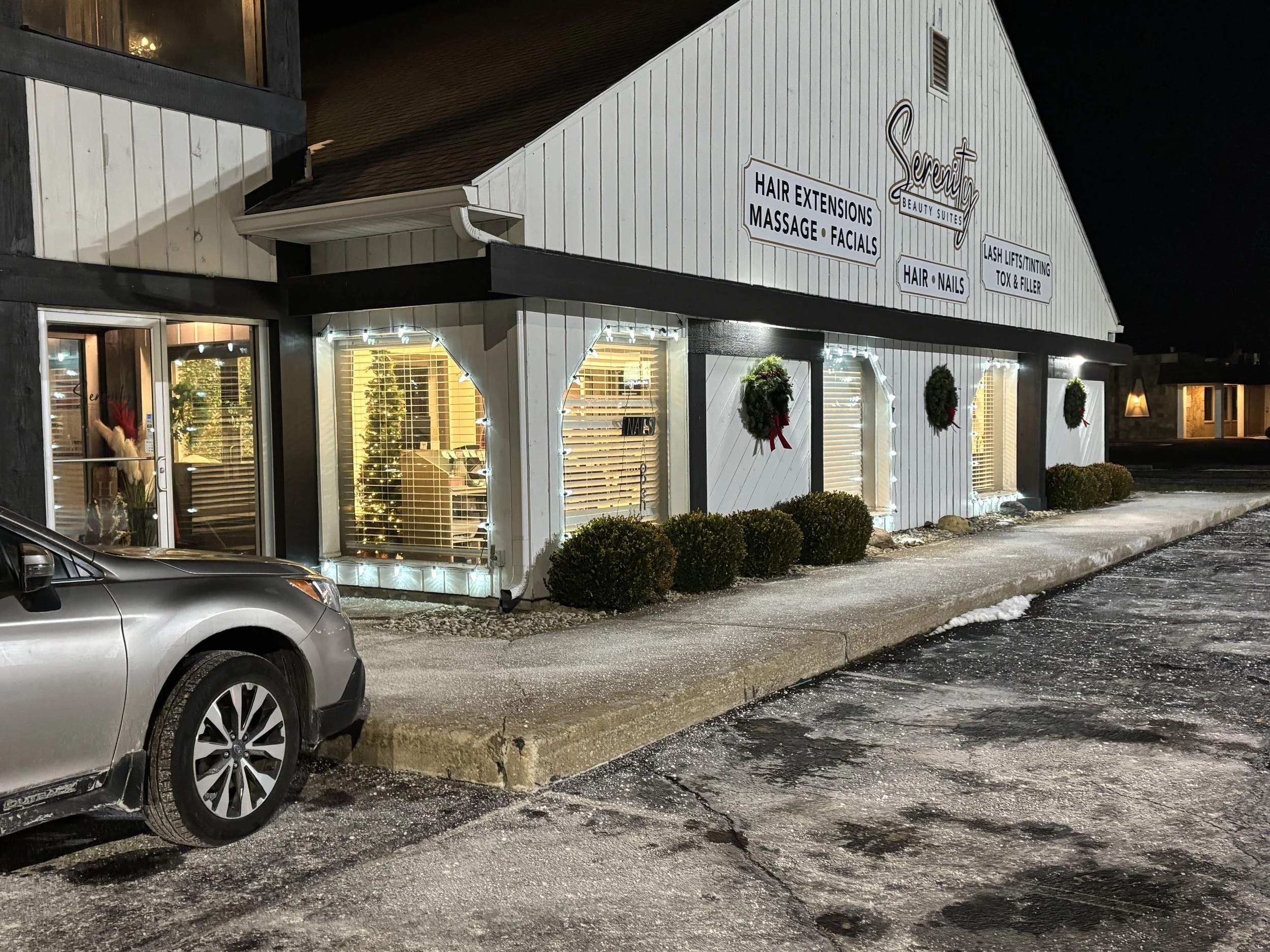Exterior view of a beauty salon at night decorated with holiday wreaths and lights, with parking lot and a silver car in the foreground.