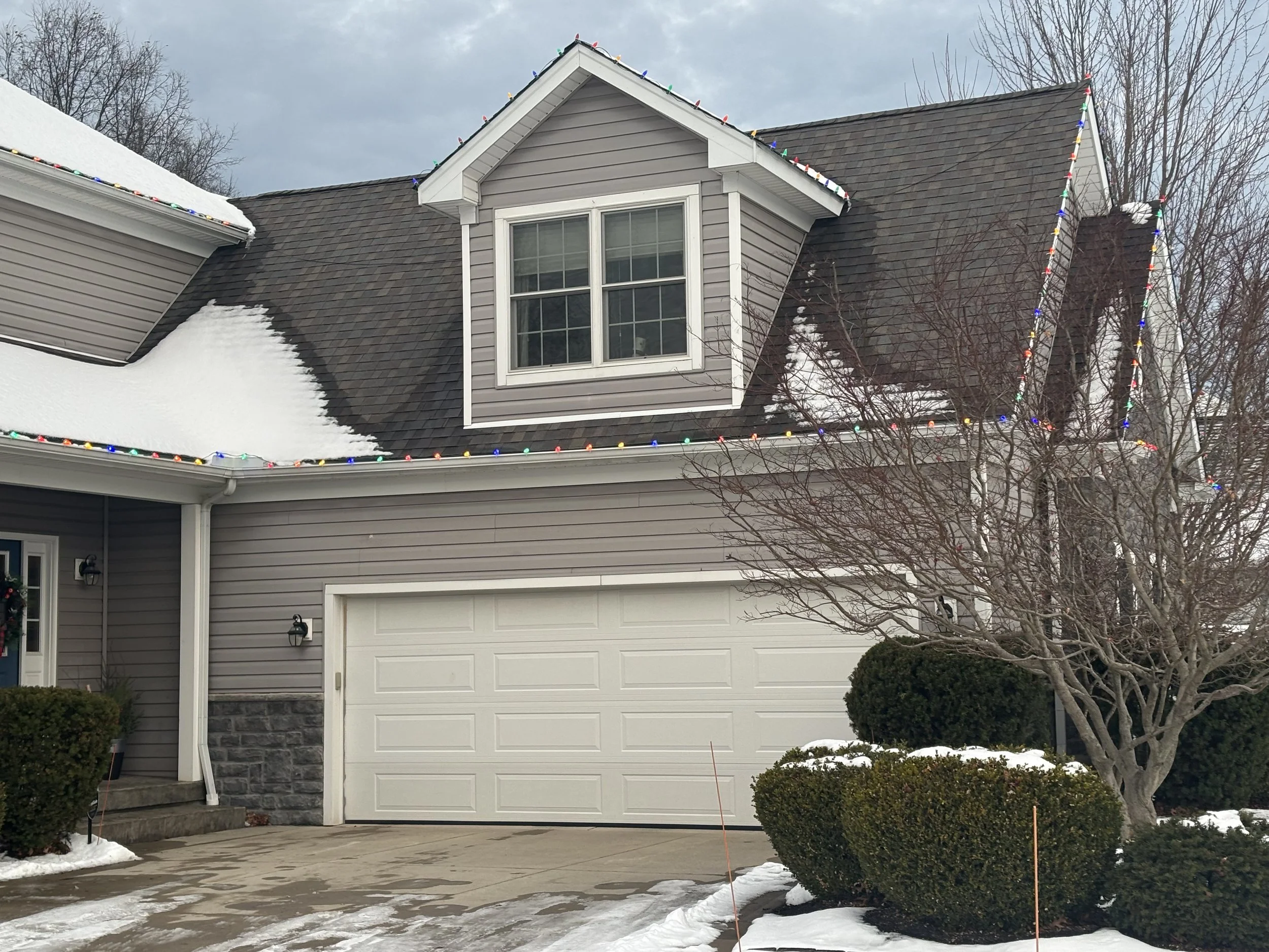 A suburban house with gray siding, a white garage door, and a dormer window. Christmas lights decorate the roof, and there is snow on the driveway, roof, and bushes in front.