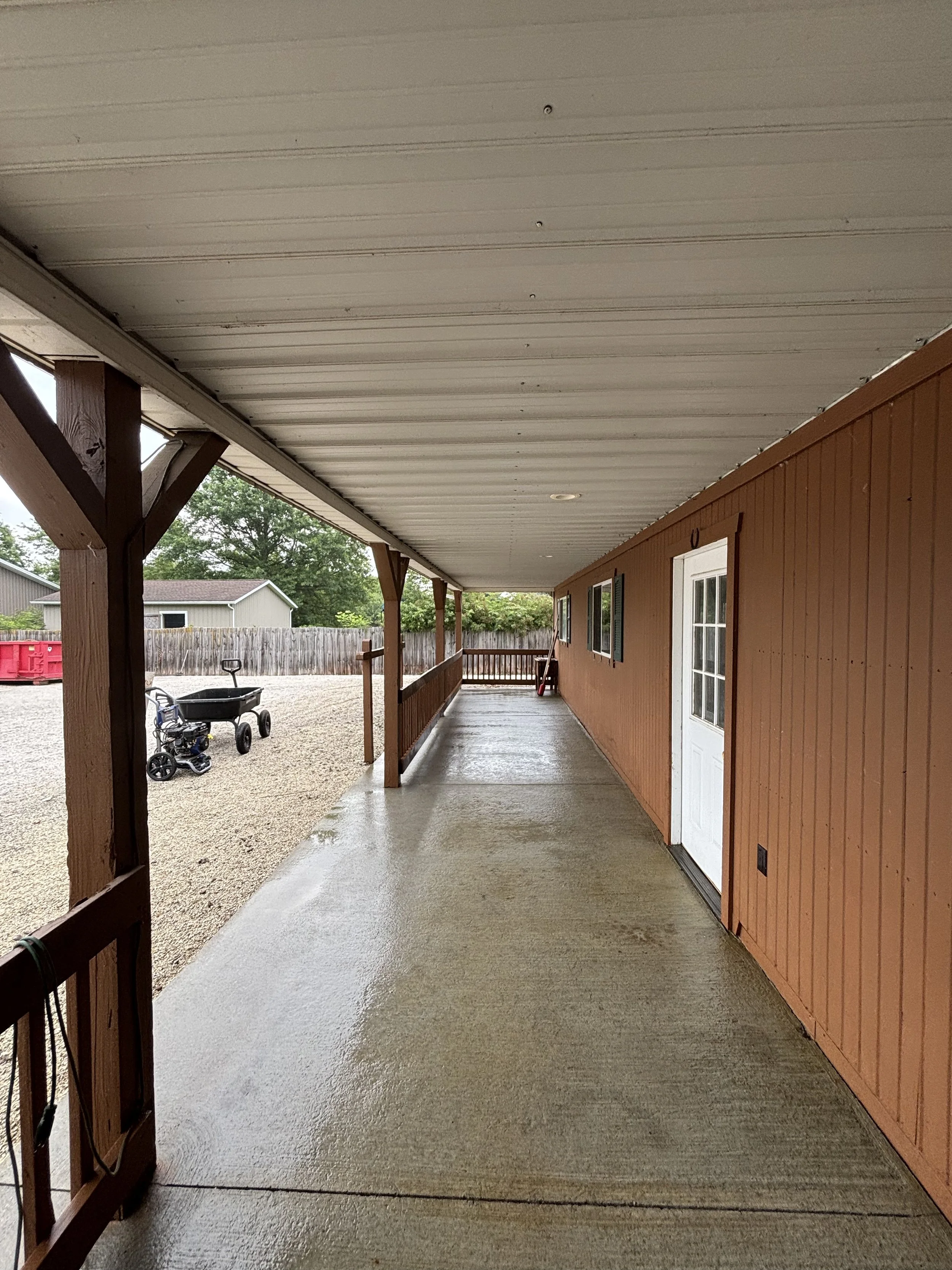 Covered porch area of a house with a concrete floor, brown wooden walls, and white ceiling. There are two windows, a white door, and outdoor toys including a wagon and children's tricycle. The yard is gravel with a wooden fence and some trees in the 