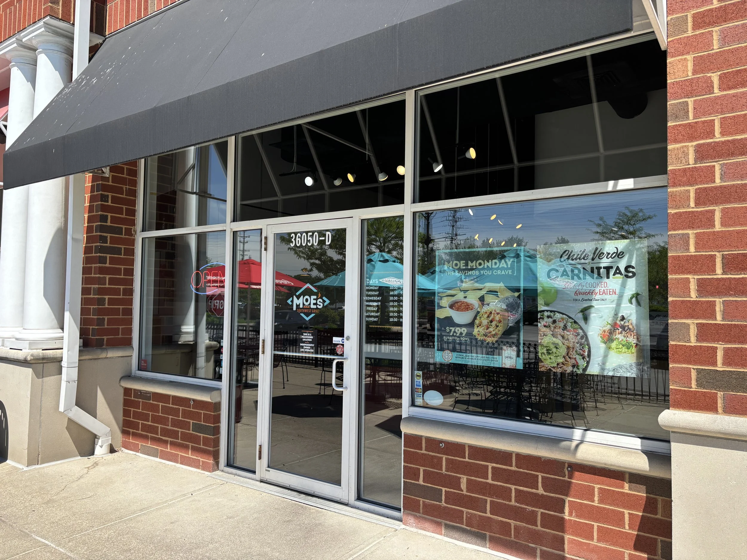 The storefront of Moe's Southwest Grill with large glass windows displaying promotional posters for food items and a neon 'Open' sign inside. The entrance door is made of glass with the address 36050-D above it. Outdoor patio tables are visible throu
