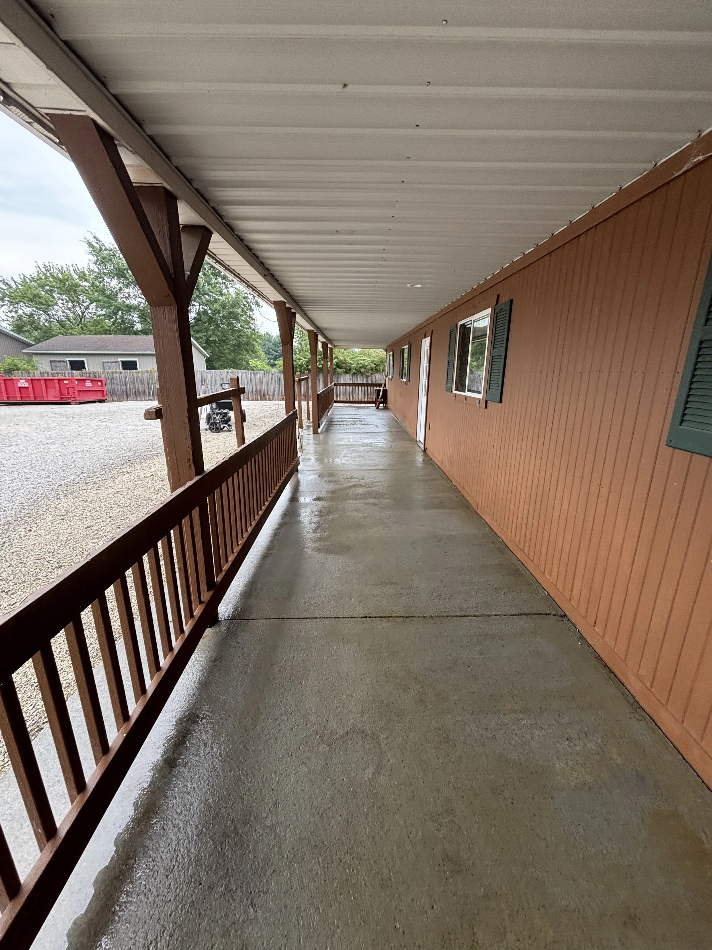 Empty covered porch with wooden railing and brown siding wall, windows with green shutters, and an overcast sky.