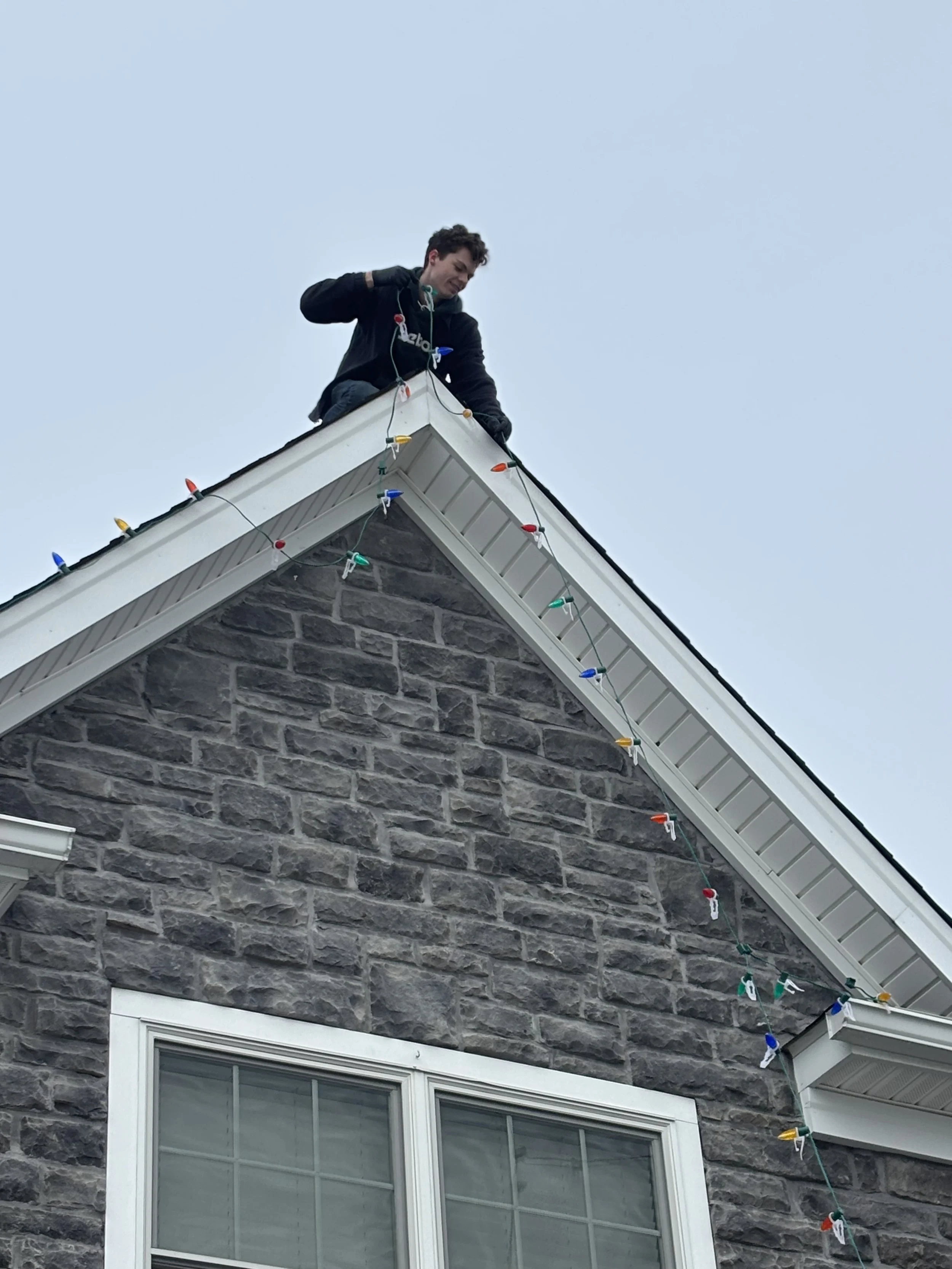 A person standing on the roof of a house, attaching Christmas lights along the edge of the roofline.