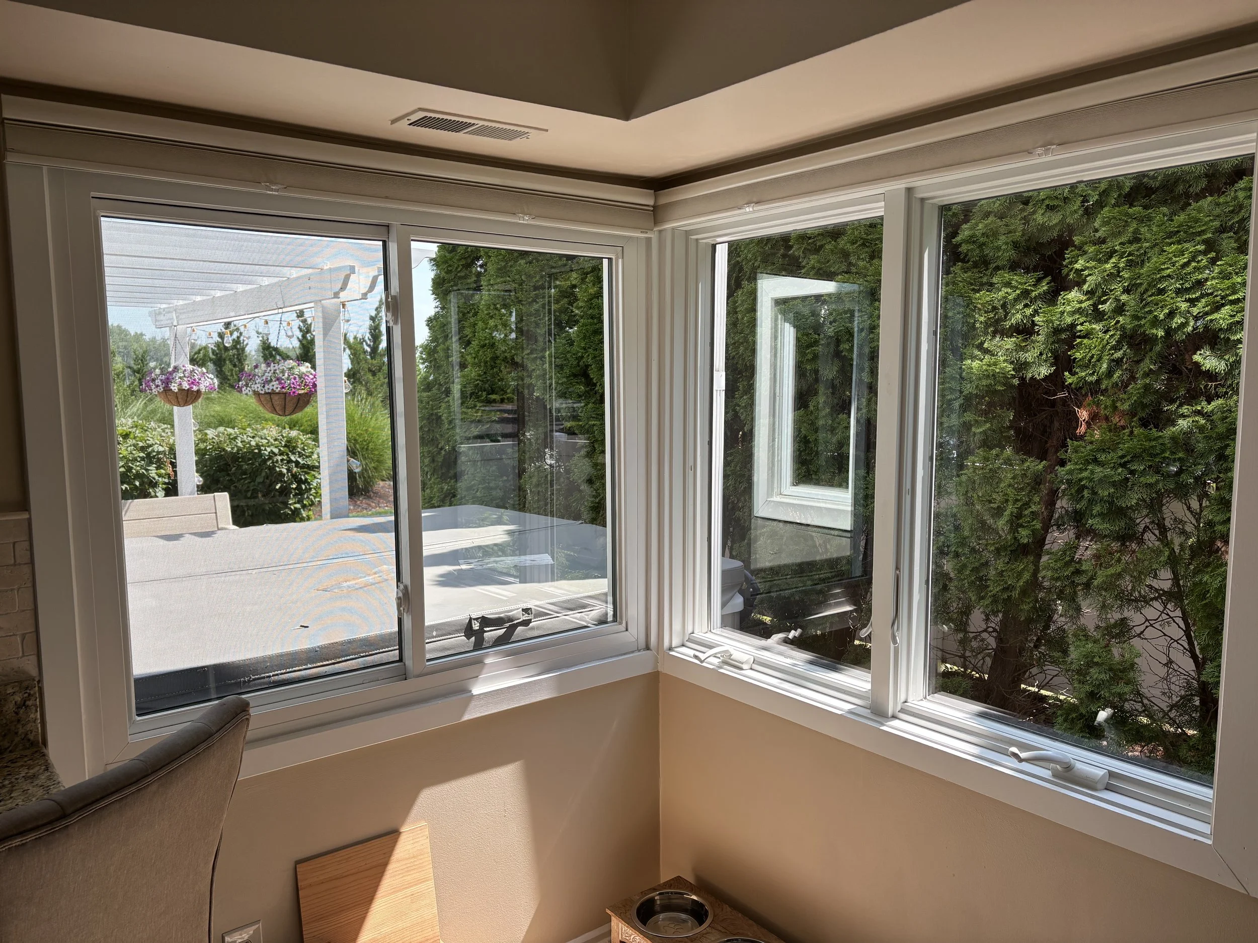 View from inside a house showing a window with a patio and garden outside, including hanging flower baskets, greenery, and a white pergola.