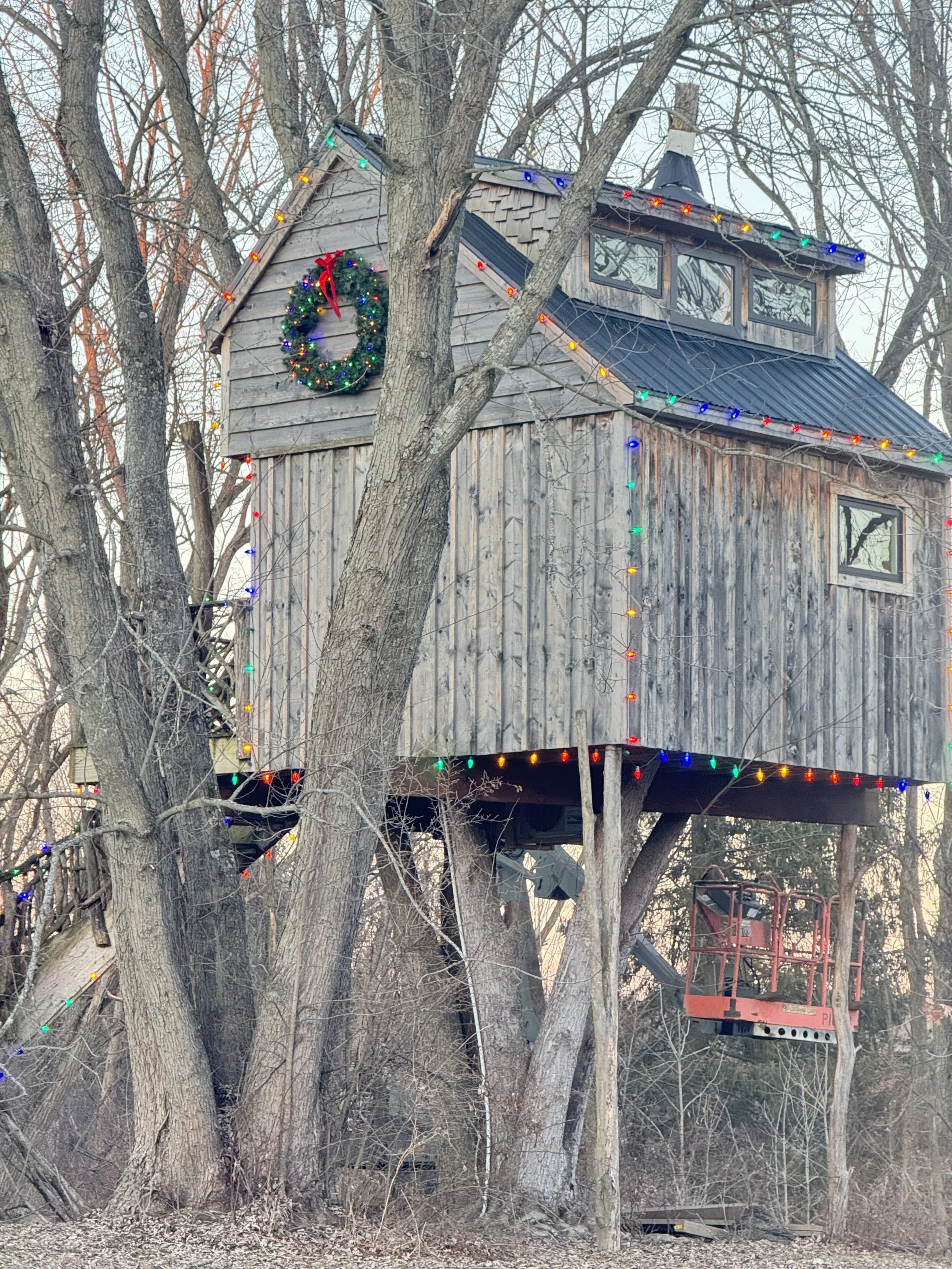 A rustic wooden treehouse decorated with colorful Christmas lights and a wreath, situated among bare trees during winter.