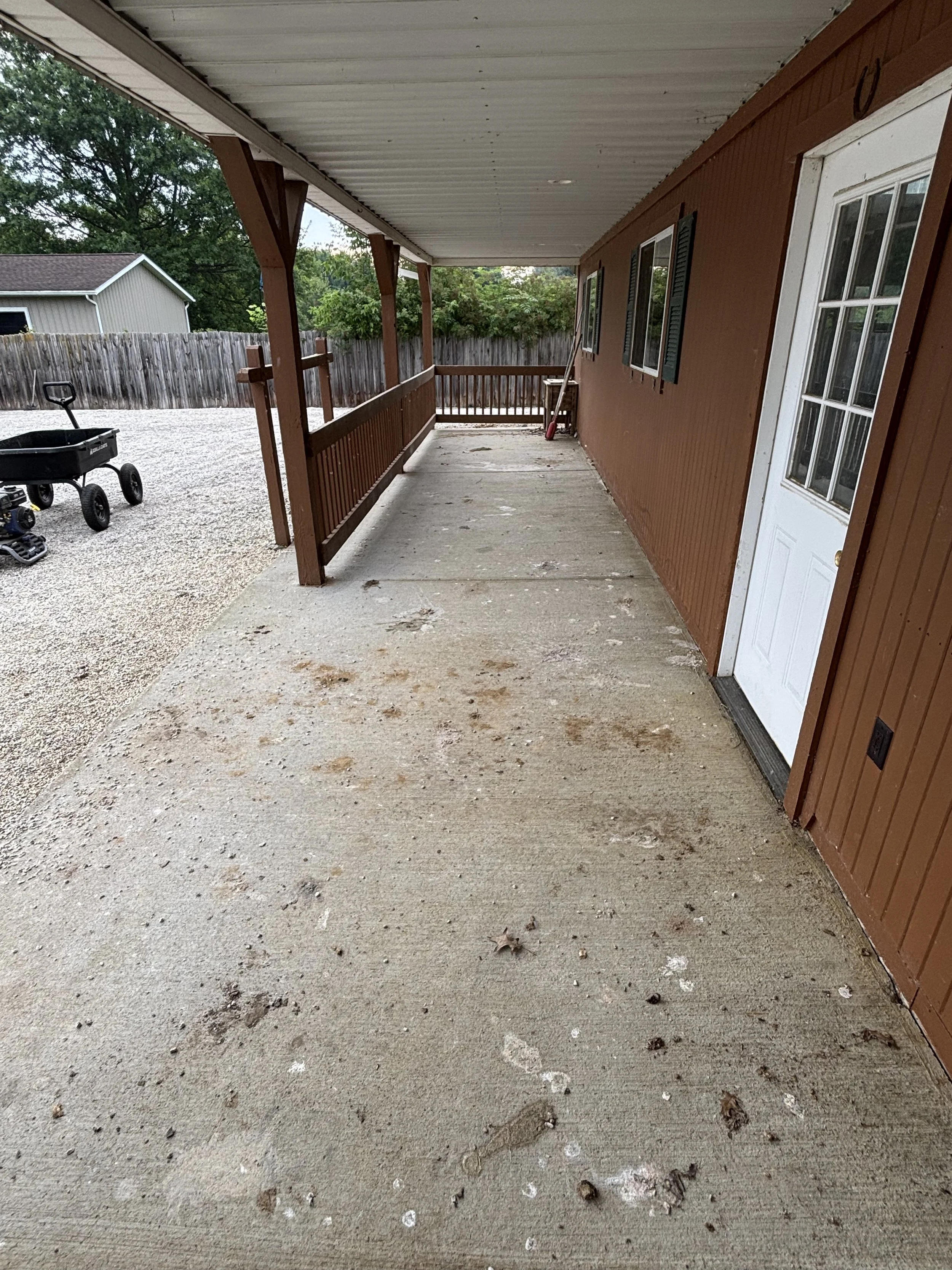 Outdoor porch area with concrete floor, brown siding, white door, and a wooden railing. There is a small wagon and some tools near the back, and the porch overlooks a gravel yard with trees and a neighboring building.