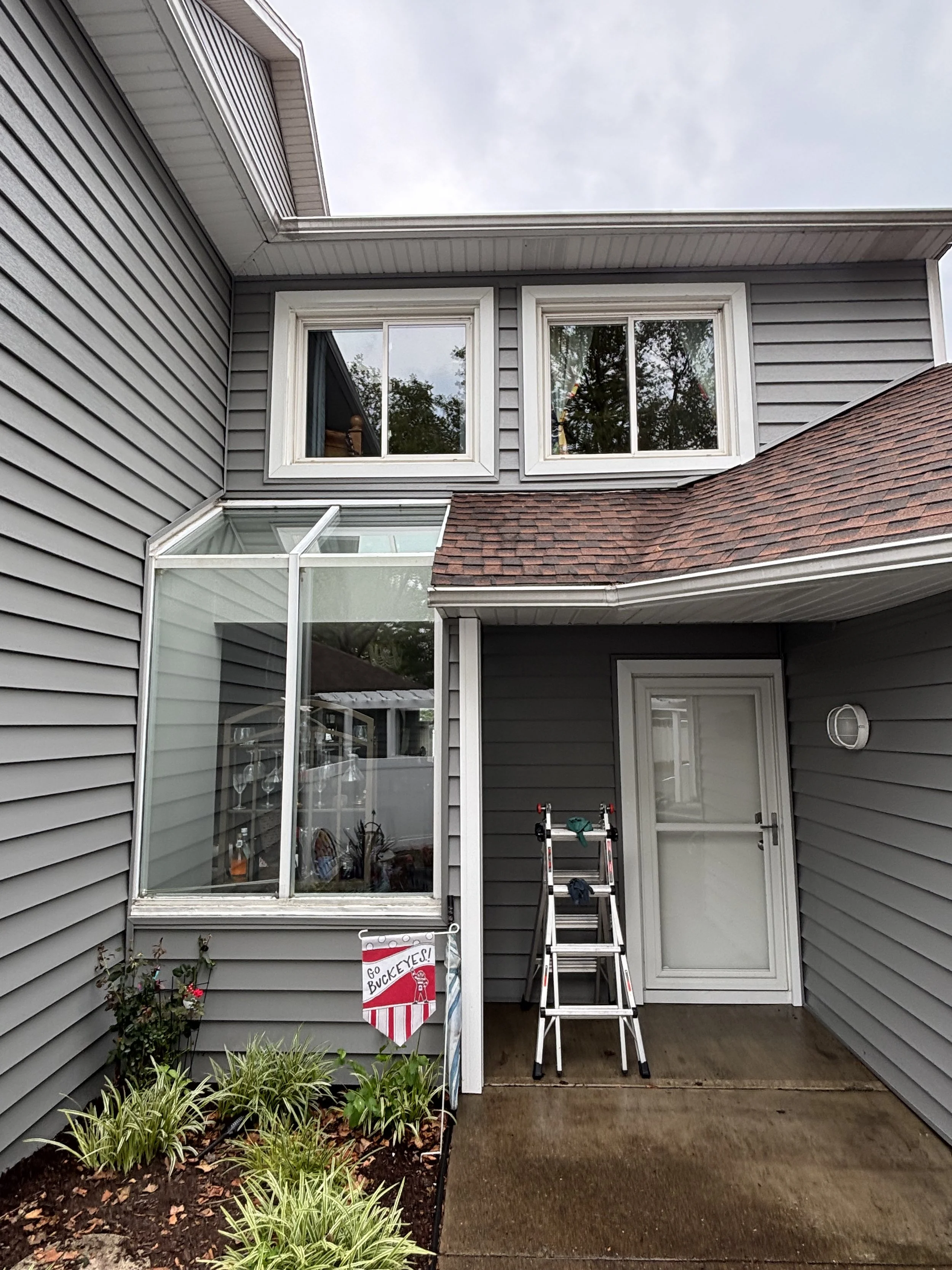 View of a gray house exterior with a small covered porch, glass-enclosed patio, a ladder, and gardening plants, with cloudy sky in background.