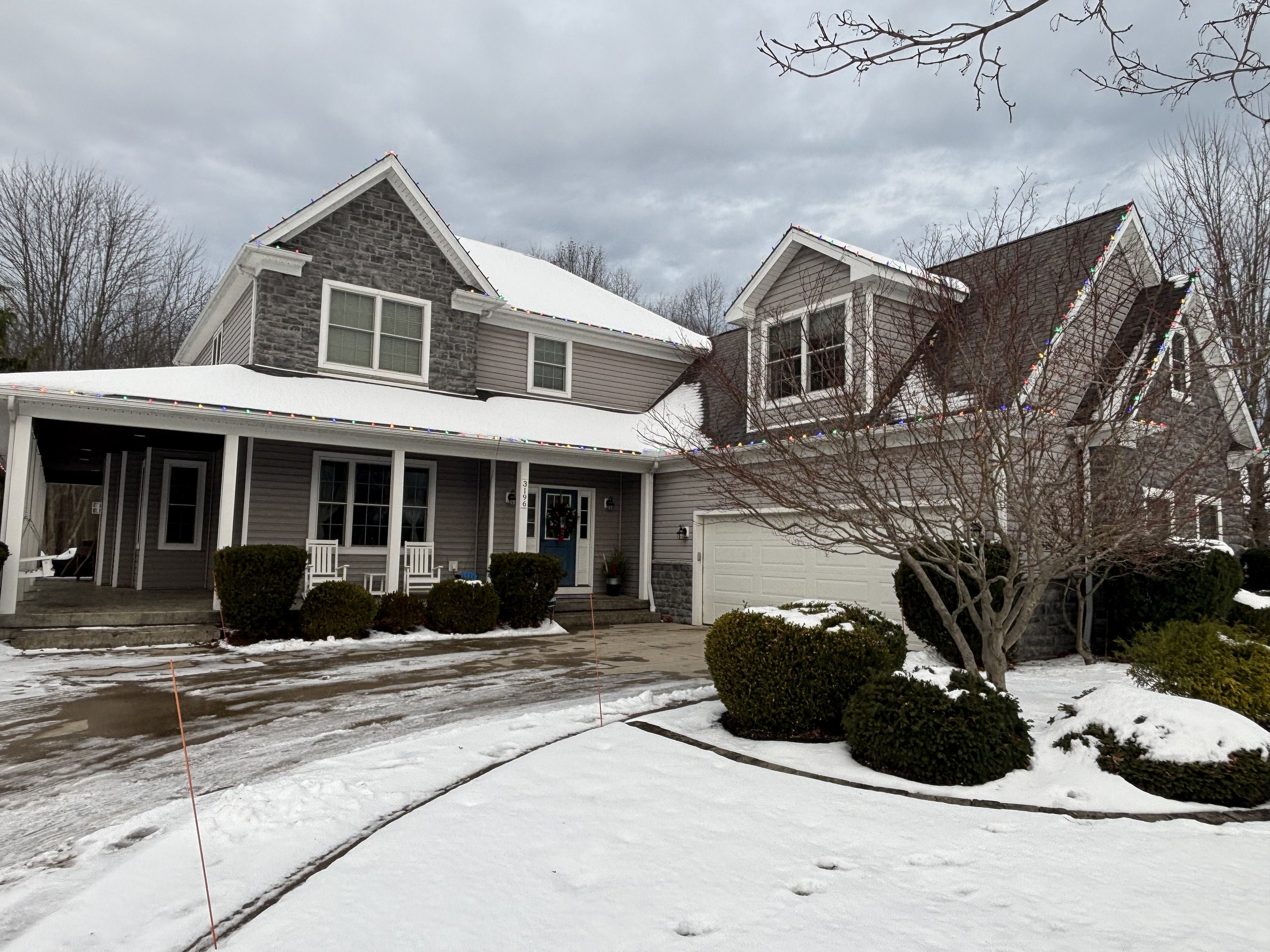A two-story house with a snow-covered yard, decorated with colorful holiday lights along the roofline. The house has a front porch with white chairs, shrubbery, and a leafless tree in the yard.