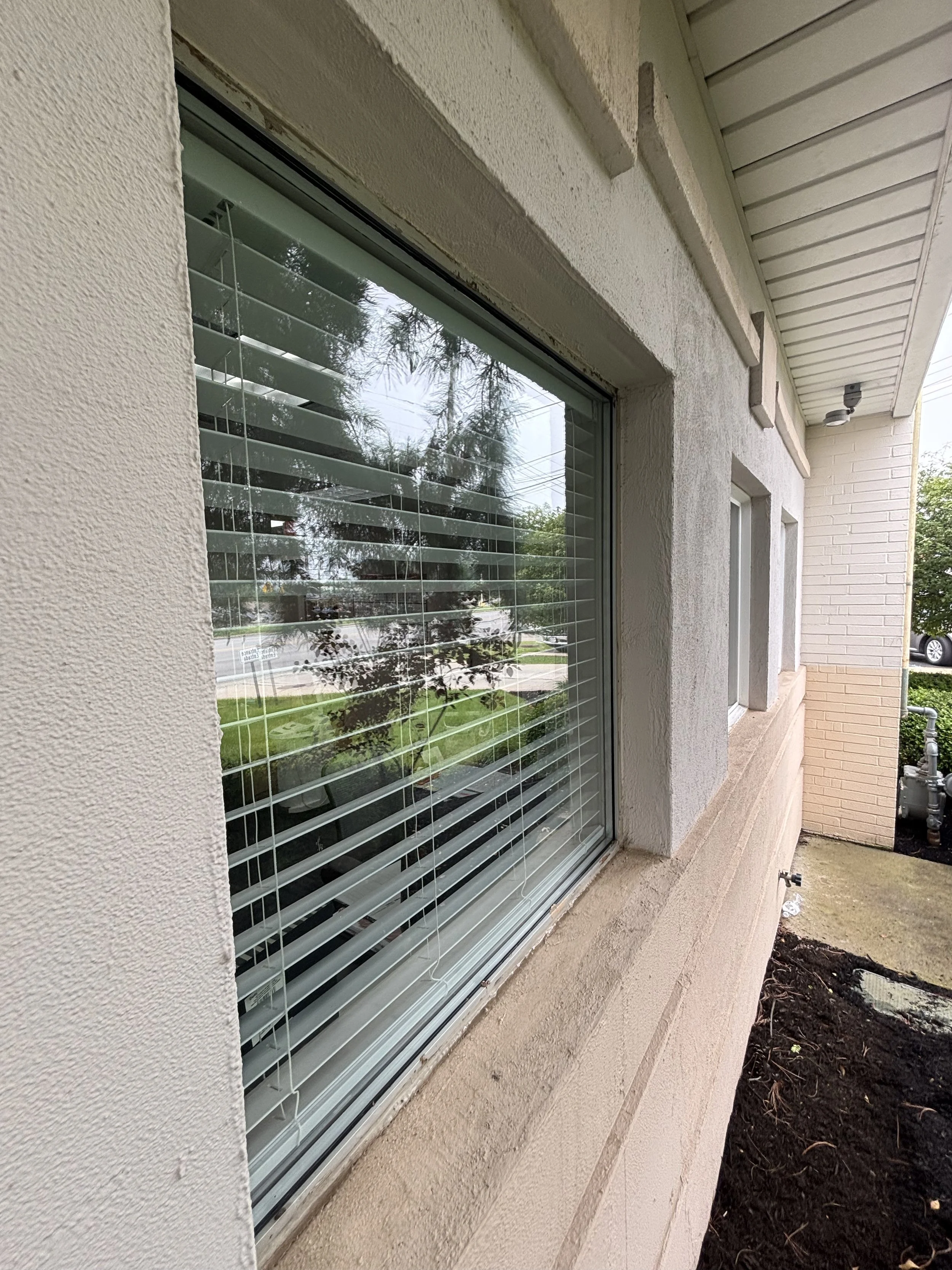 Close-up of a large window with white horizontal blinds on a beige textured house exterior, reflecting trees and a street in the glass.