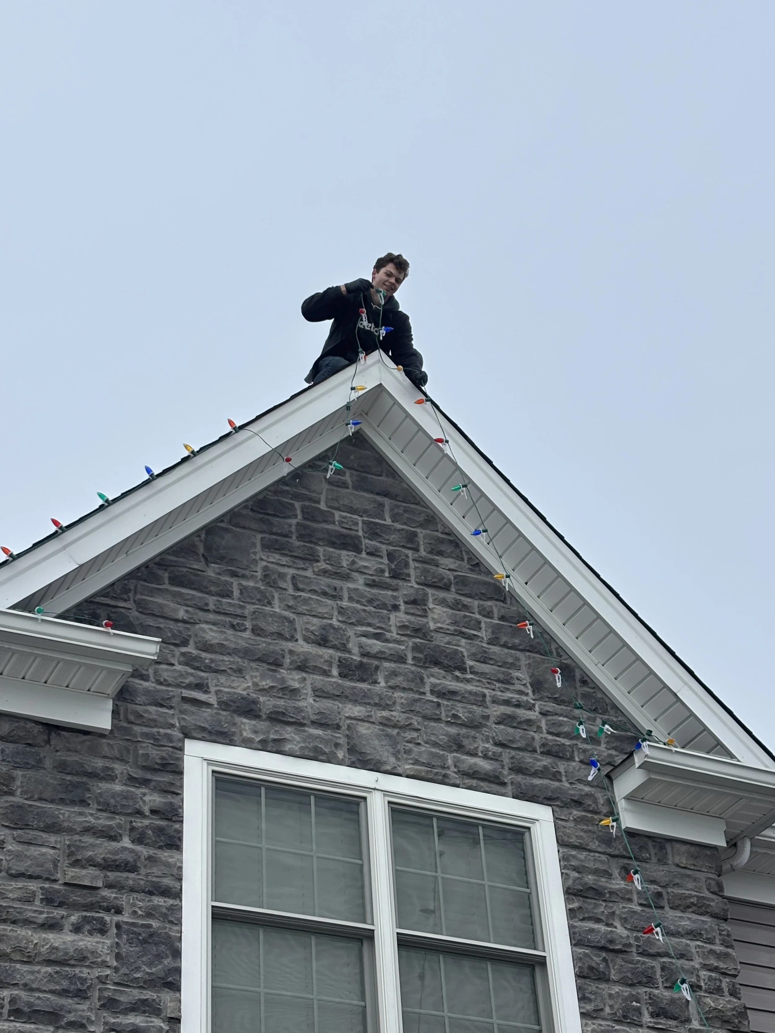 A person standing on the roof of a house decorated with colorful Christmas lights, smiling and holding a string of lights.