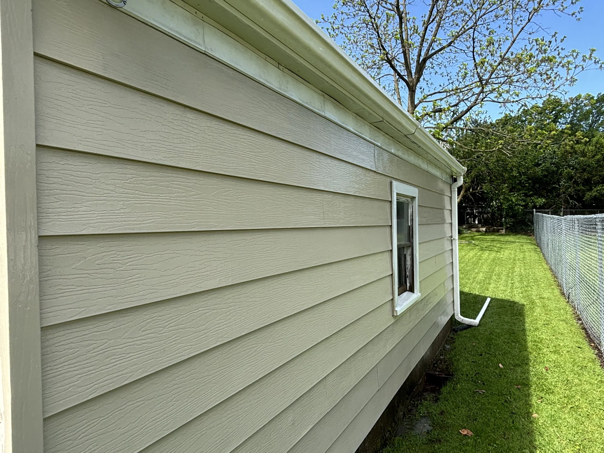 Side view of a beige house with horizontal siding, a small window, and white gutters, next to a chain-link fence and a grassy yard with trees.