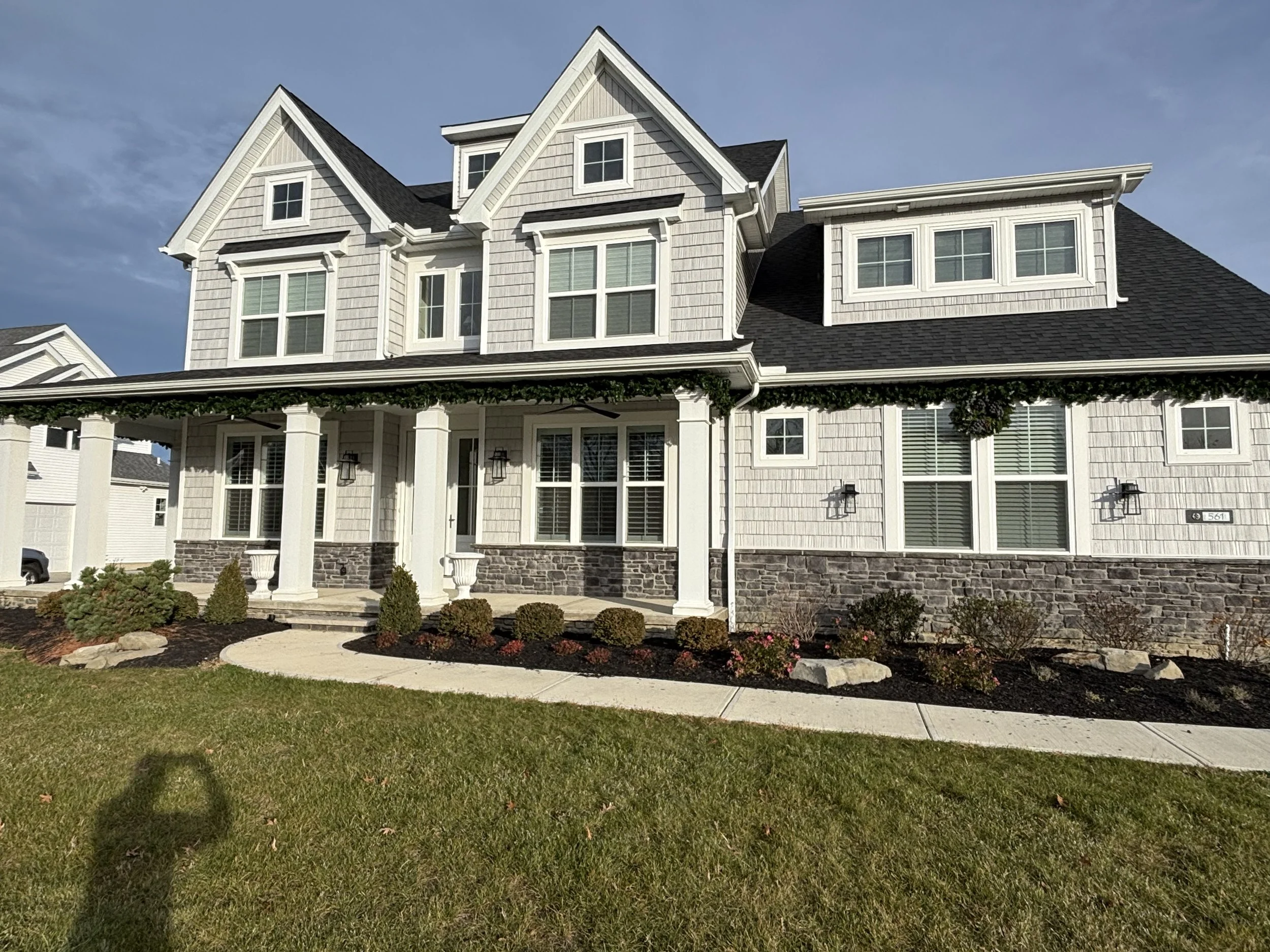 Large multi-story house with a white exterior, black roof, multiple windows, and front porch with columns, surrounded by a landscaped yard with shrubs and a sidewalk.