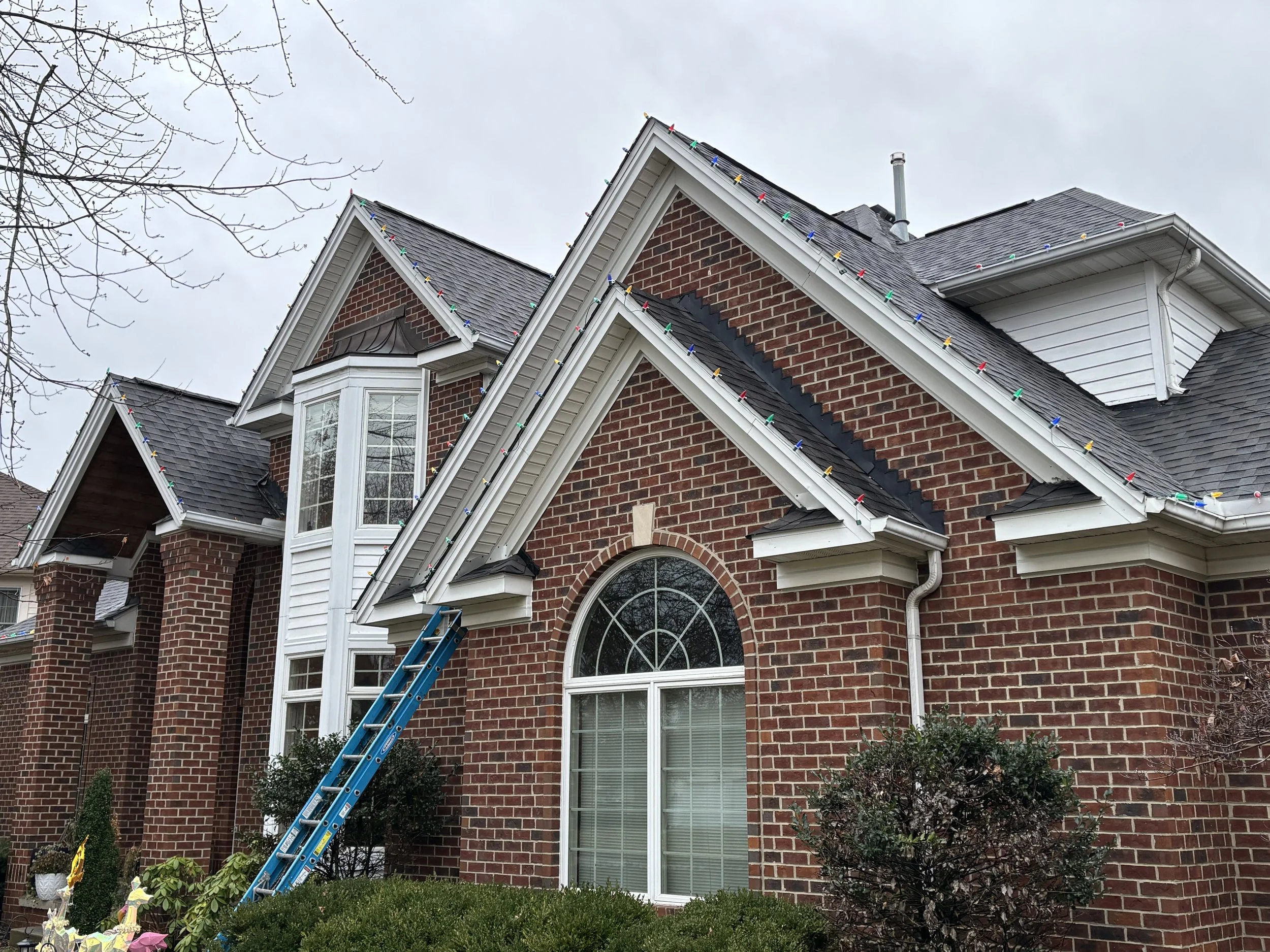 Decorated brick house with Christmas lights on roof, ladder leaning against the house, snow on the ground, bare trees, large arched window, and shrubs in the yard.